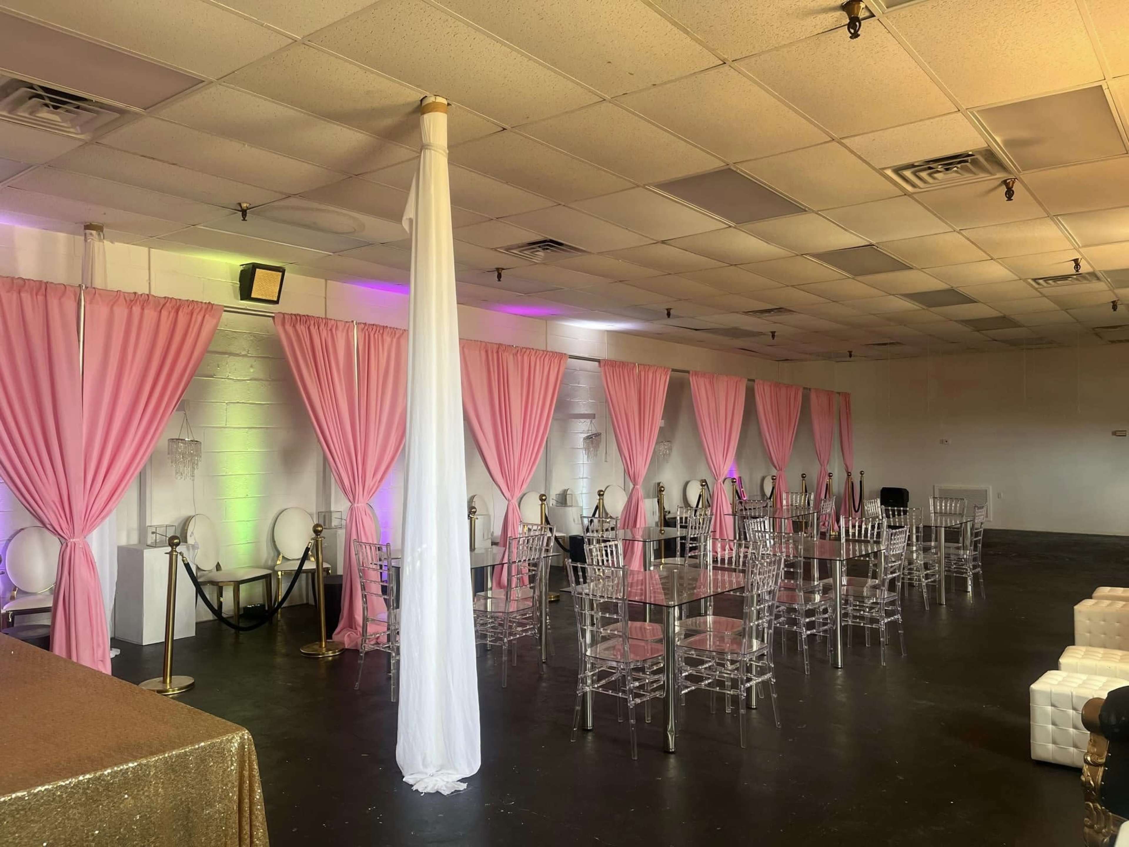 An event space with pink drapes, clear acrylic chairs, and a gold tablecloth, arranged for a gathering.