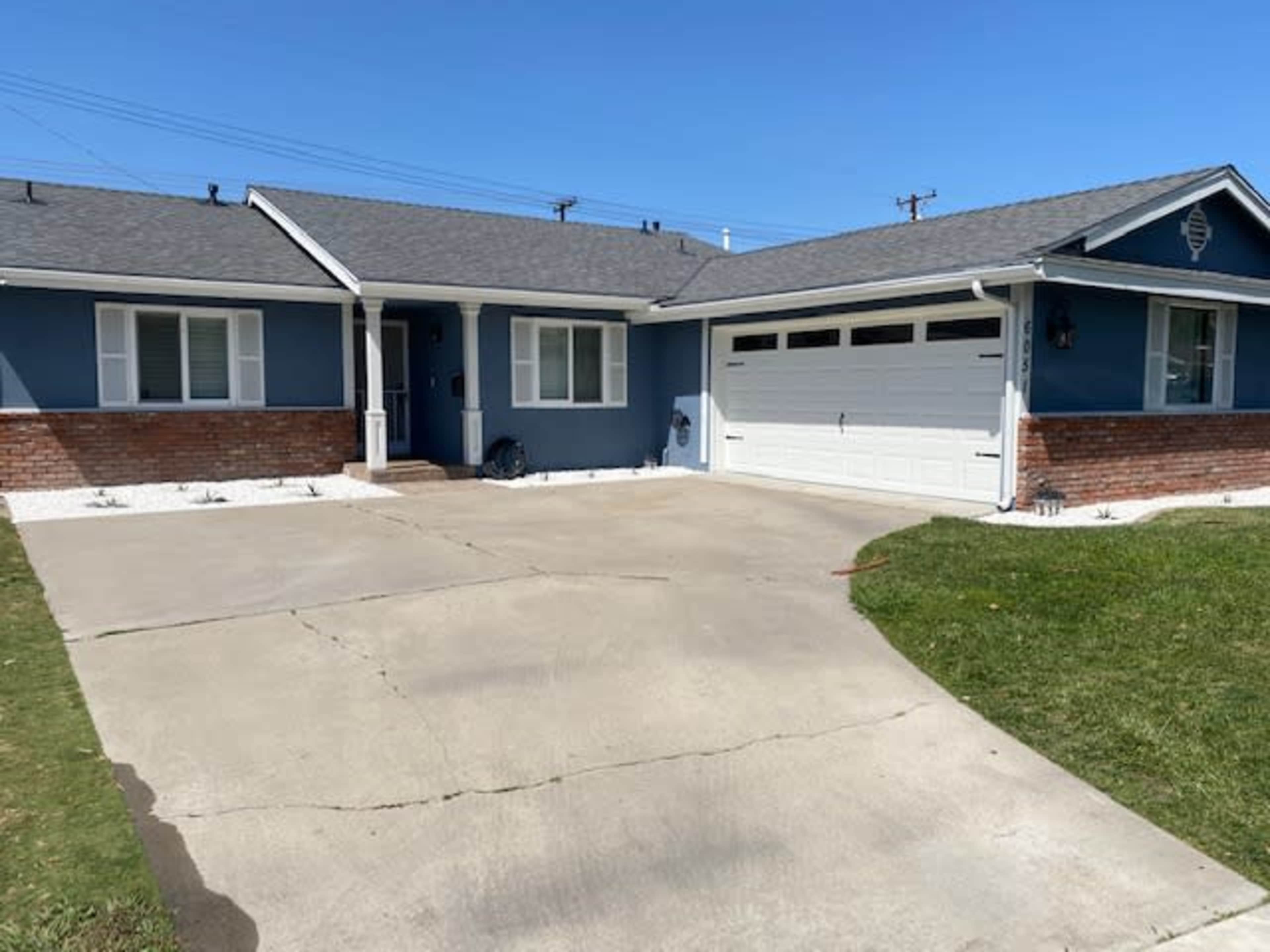 A single-story blue house with a brick facade has a concrete driveway leading to a garage on a clear day.