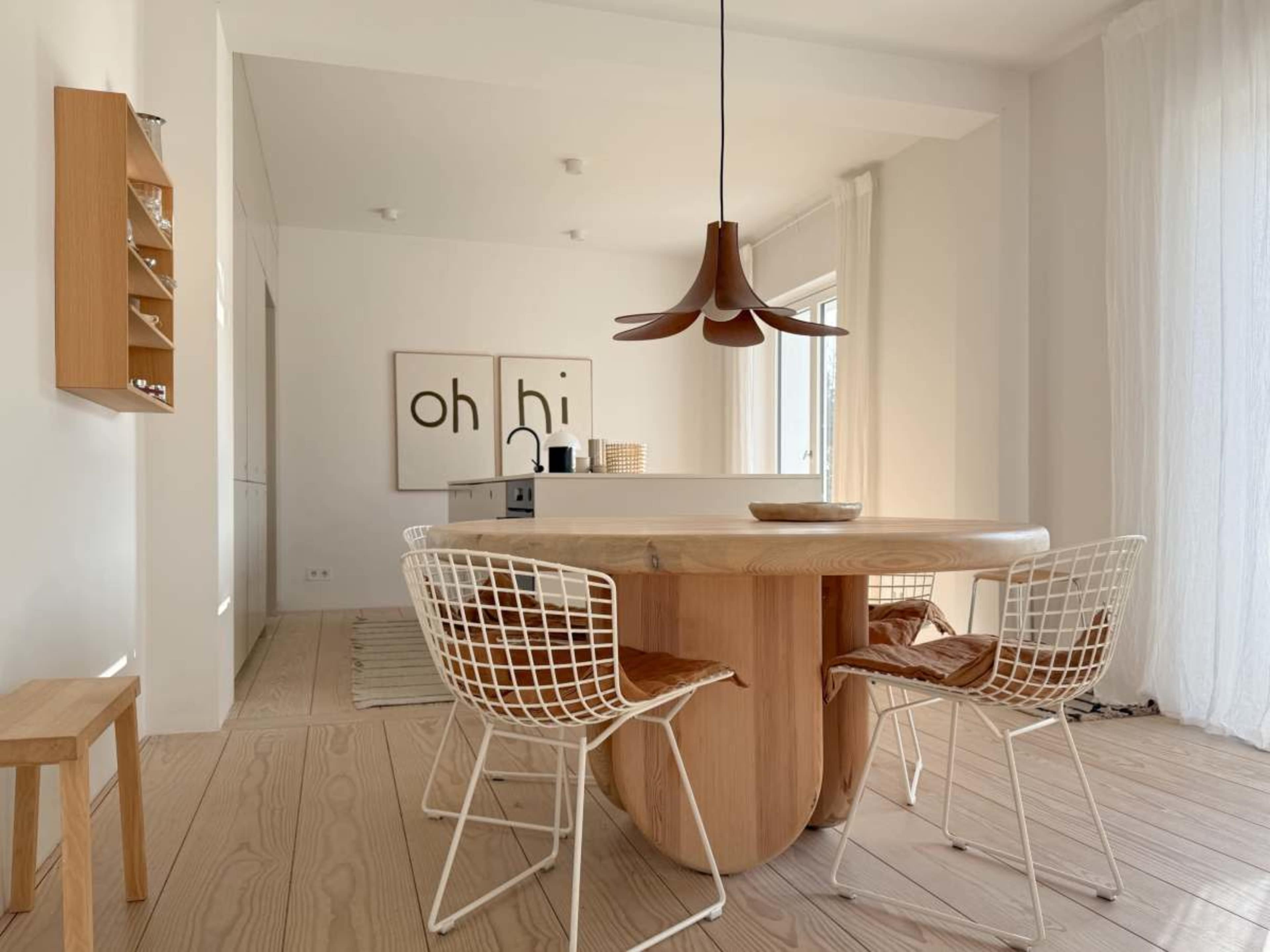 The image shows a modern dining area featuring a round wooden table with a distinctive base, accompanied by four wireframe chairs, and a minimalist kitchen visible in the background.