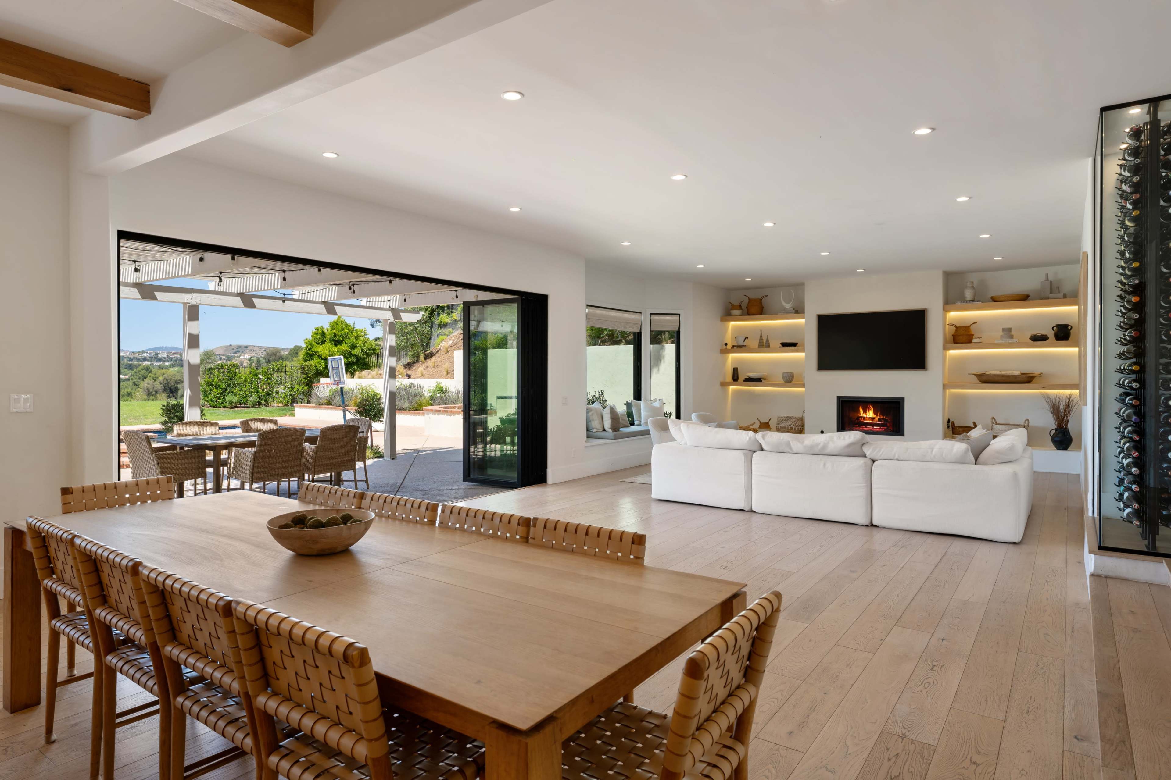 A modern interior showcases a dining area with a wooden table and woven chairs, adjacent to a living room featuring a fireplace and large glass doors leading to an outdoor patio.