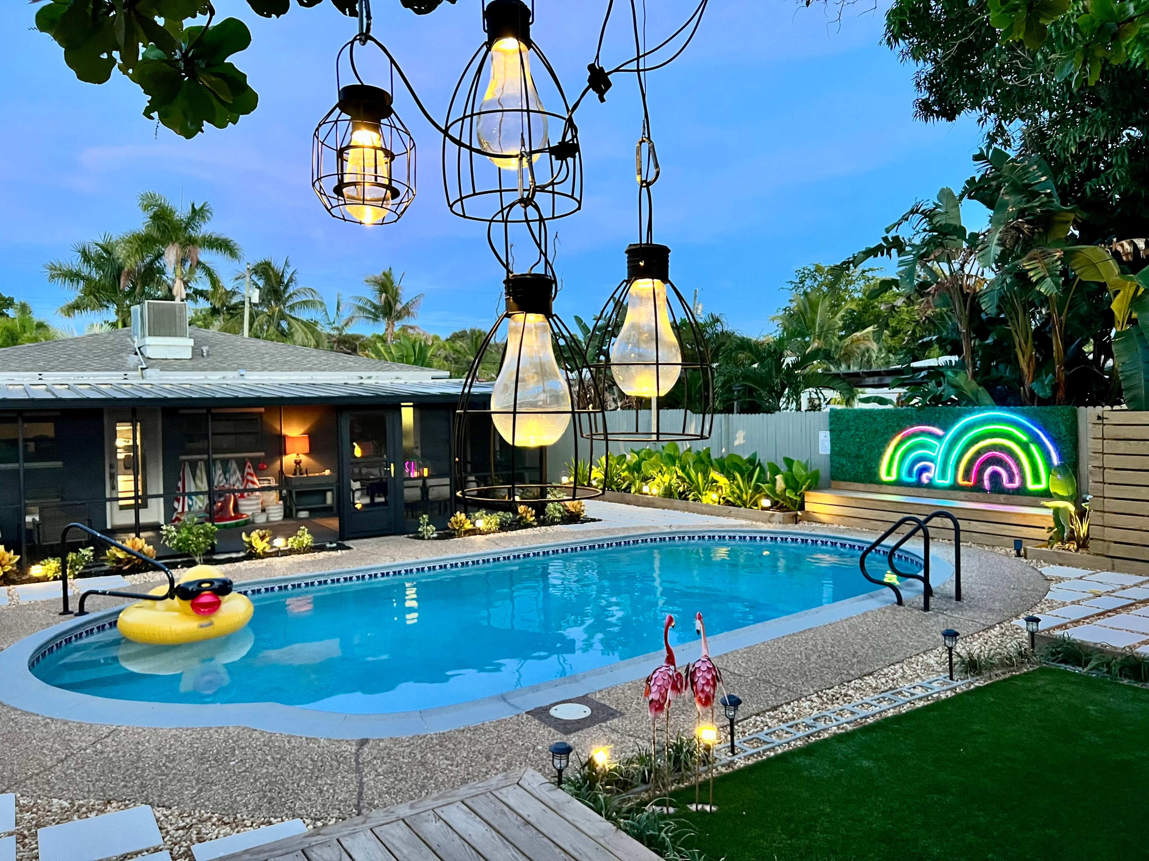 The image shows a backyard with a swimming pool surrounded by tropical plants, decorative lights hanging above, and a colorful light display on the wall.