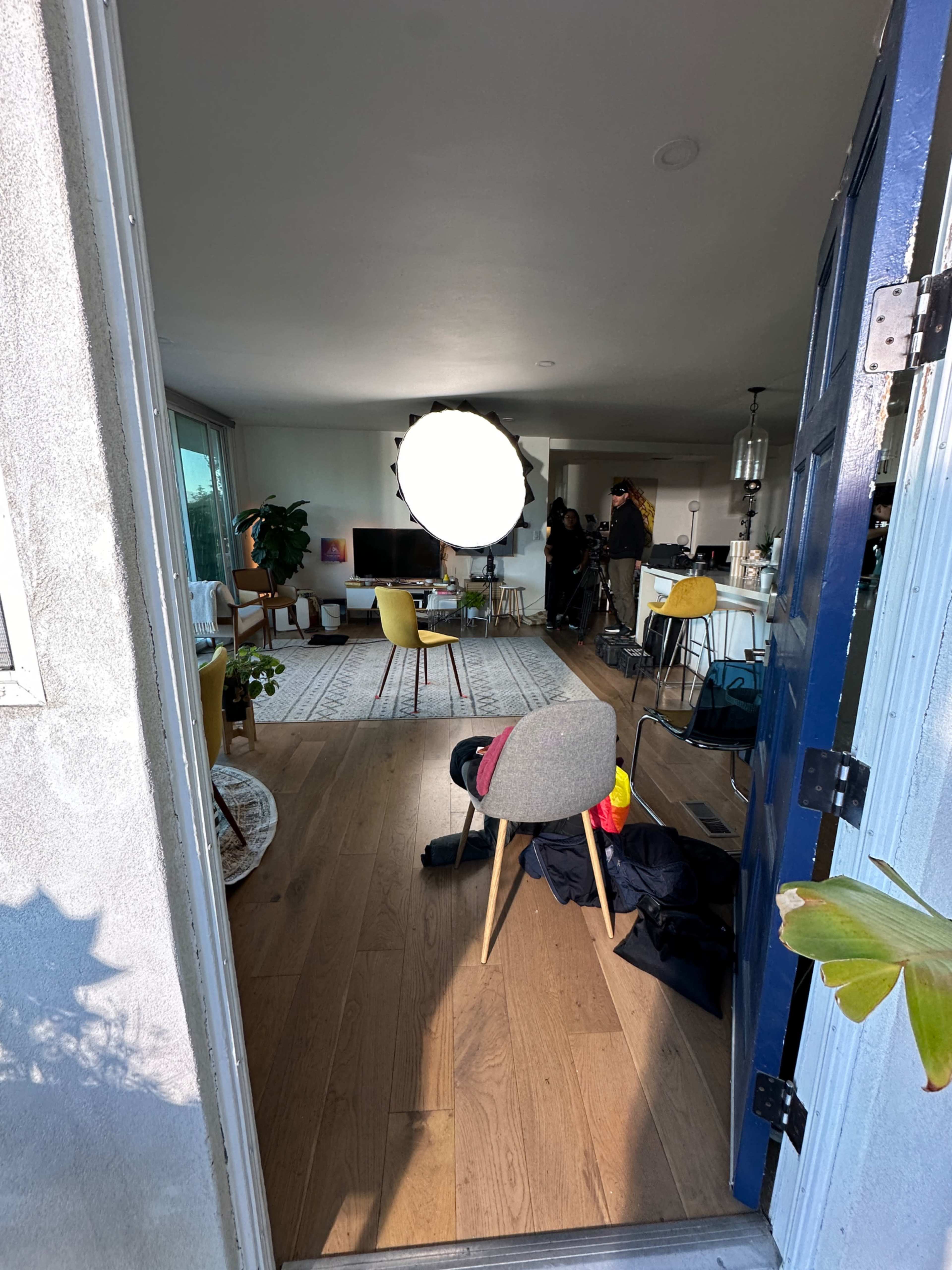 A brightly lit living room is visible through an open door, with a film crew set up and a large circular light fixture in the center.