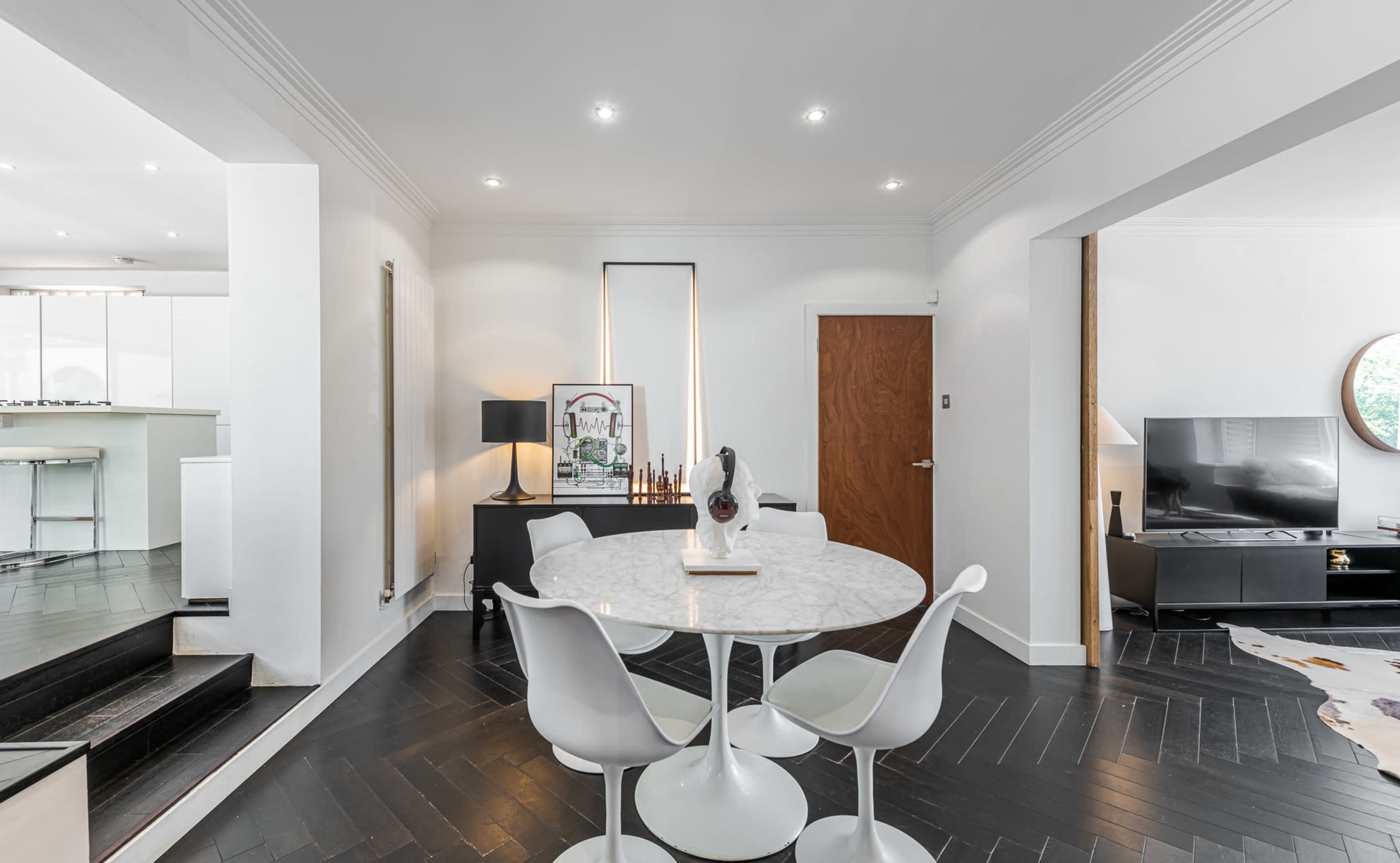 A modern interior features a round dining table with white chairs, a lamp, and a television set against a sleek black floor and white walls.