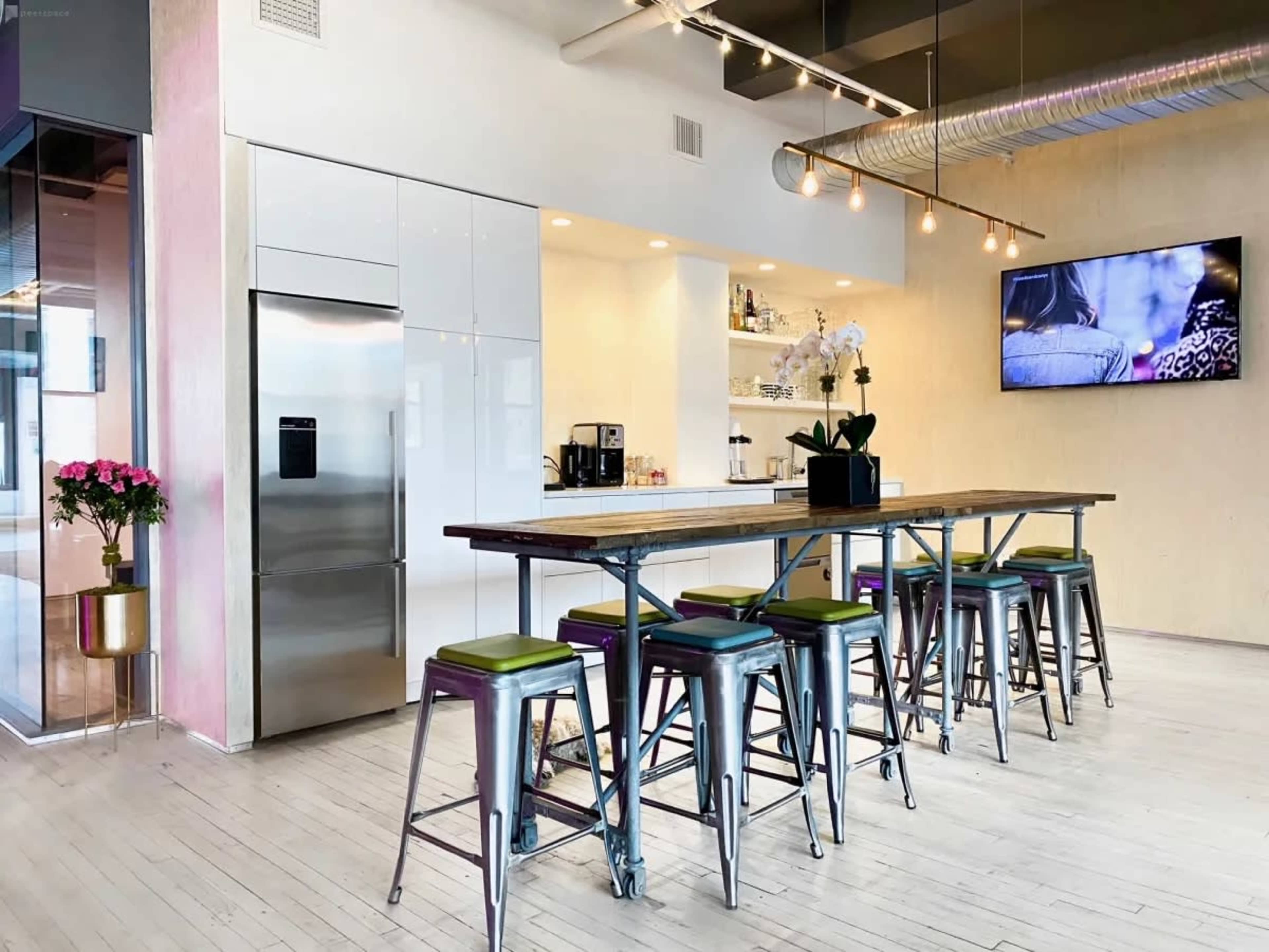 A modern kitchen area features a long wooden table with metal stools, a stainless steel refrigerator, and a wall-mounted television.