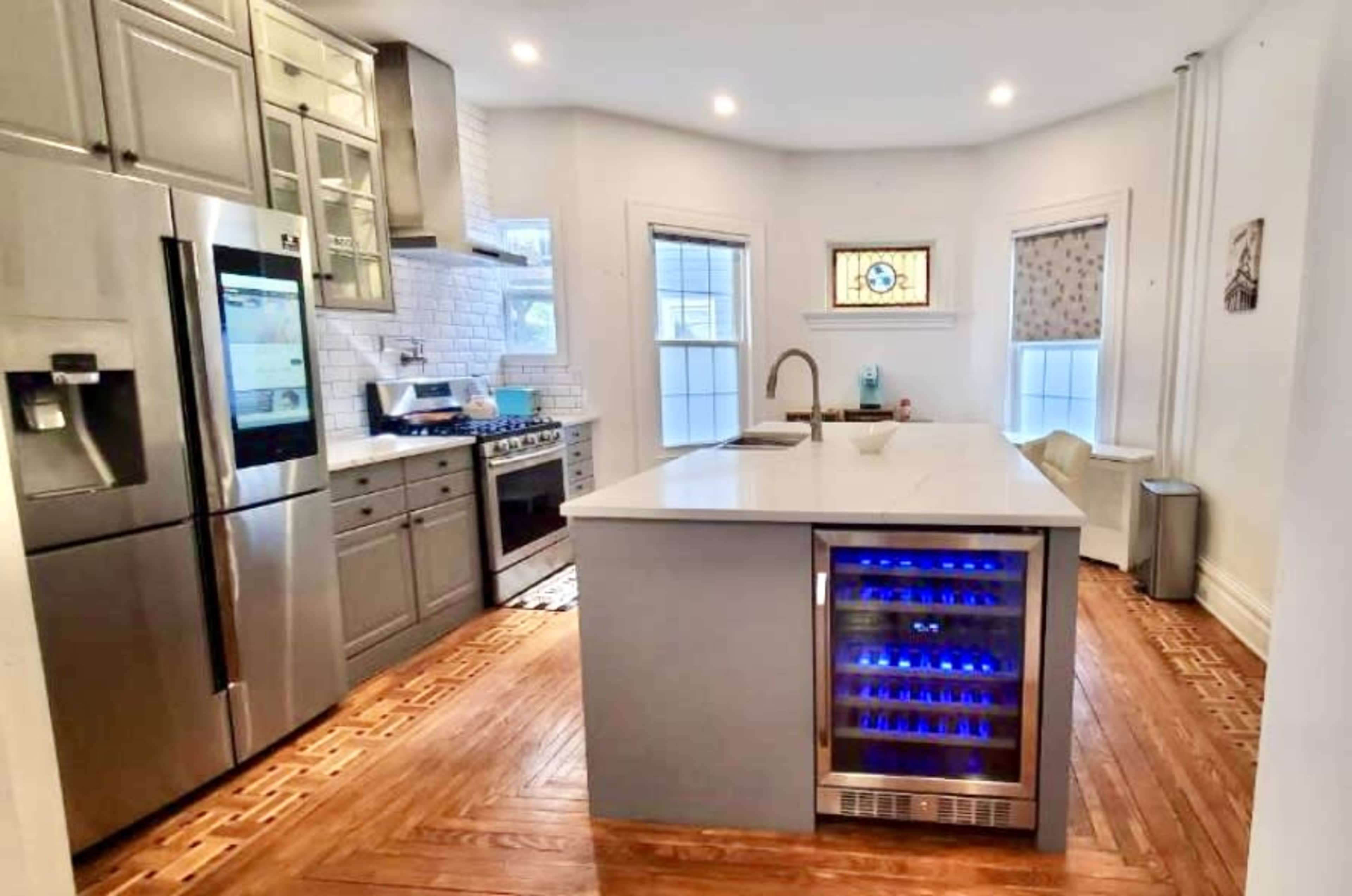 The image shows a modern kitchen with stainless steel appliances, a quartz island with a built-in wine cooler, and wooden flooring.
