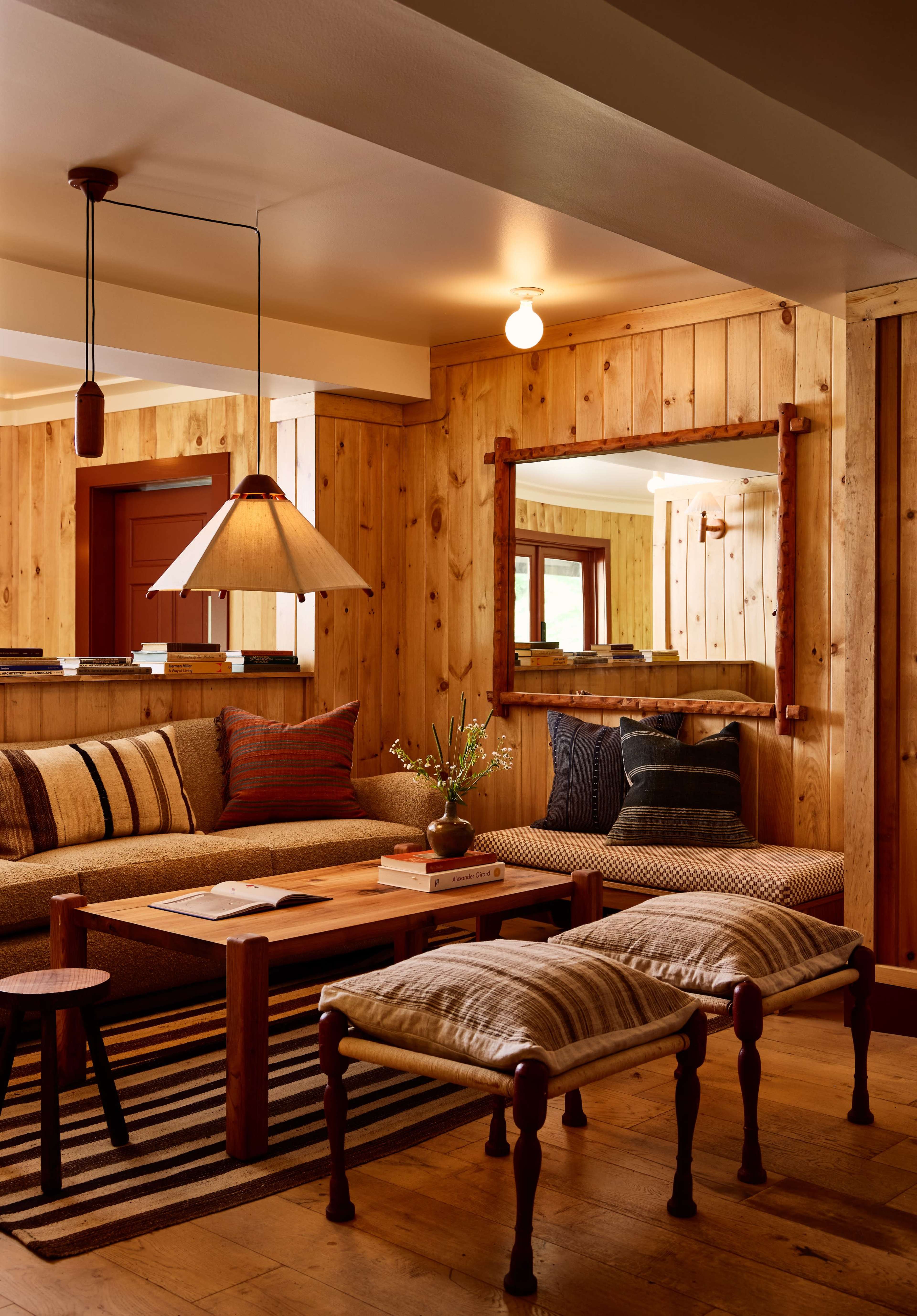 A cozy living room features wooden walls, a couch with various cushions, a lamp hanging above, and a coffee table surrounded by textile-covered stools.