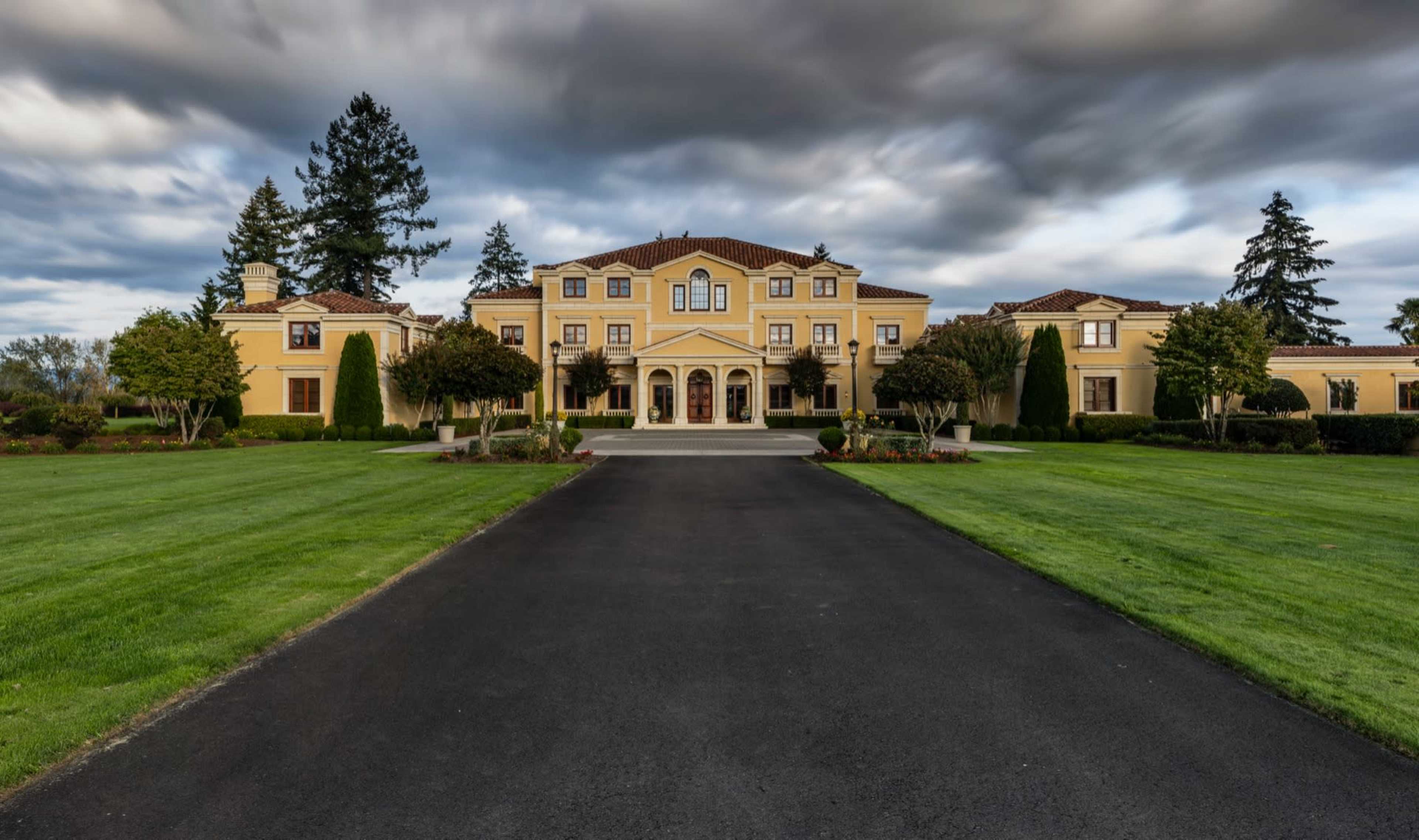 A large, yellow mansion with a tiled roof is set on manicured lawns and accessed by a long, paved driveway.