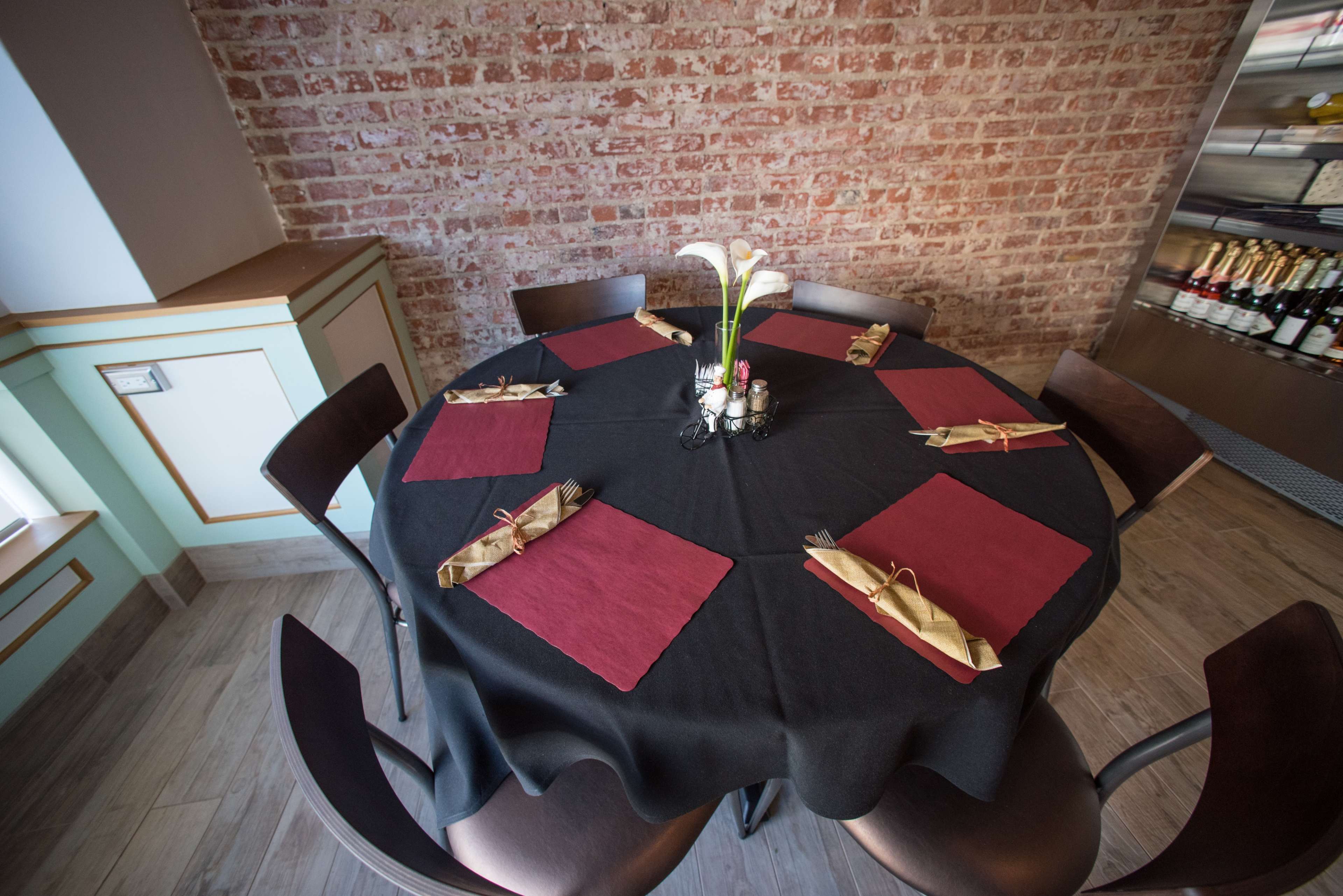 A round table is set with maroon placemats and utensils, surrounded by wooden chairs in a restaurant with a brick wall.