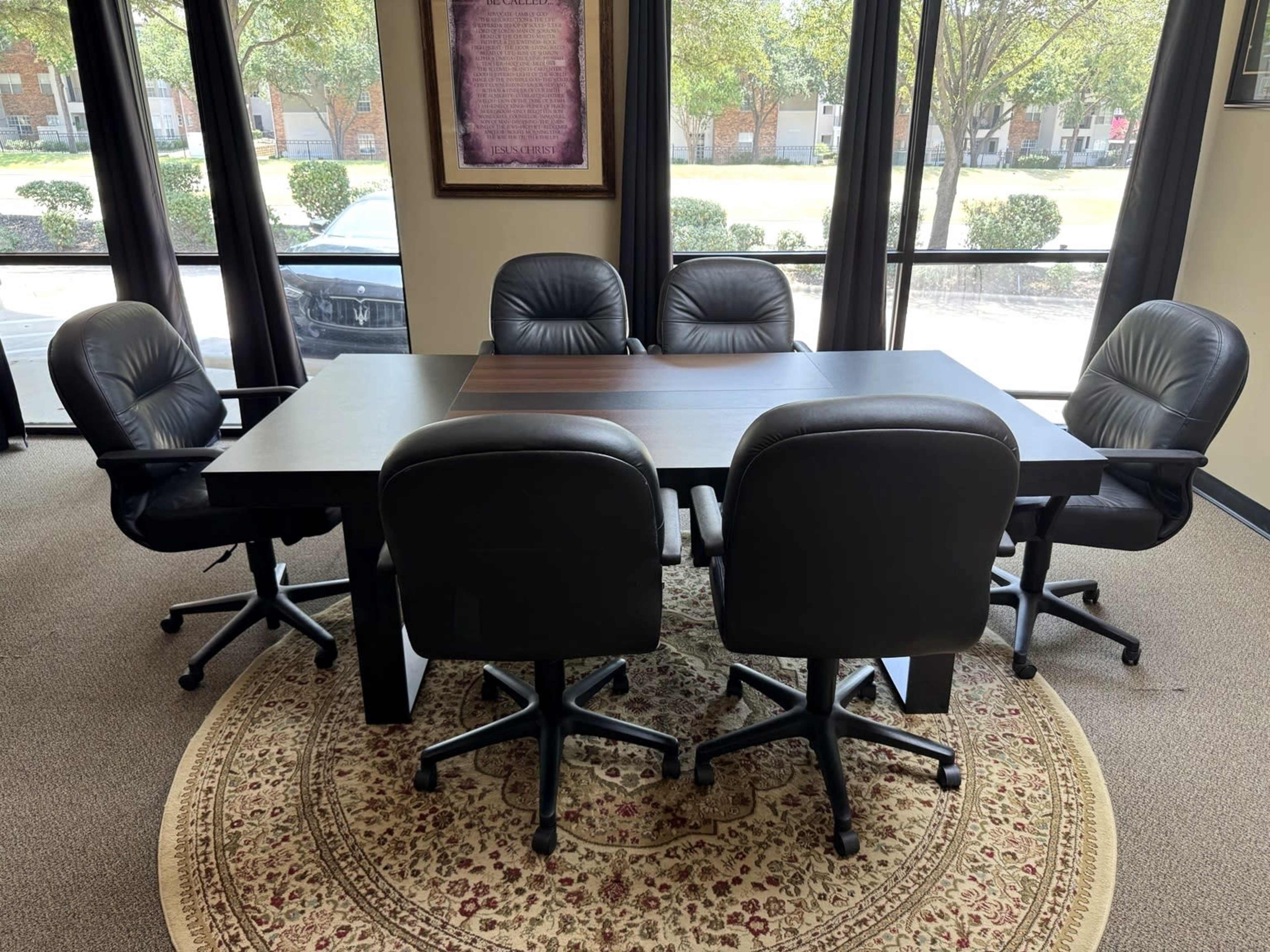 A rectangular conference table is surrounded by six black rolling chairs in an office setting, with large windows providing natural light.