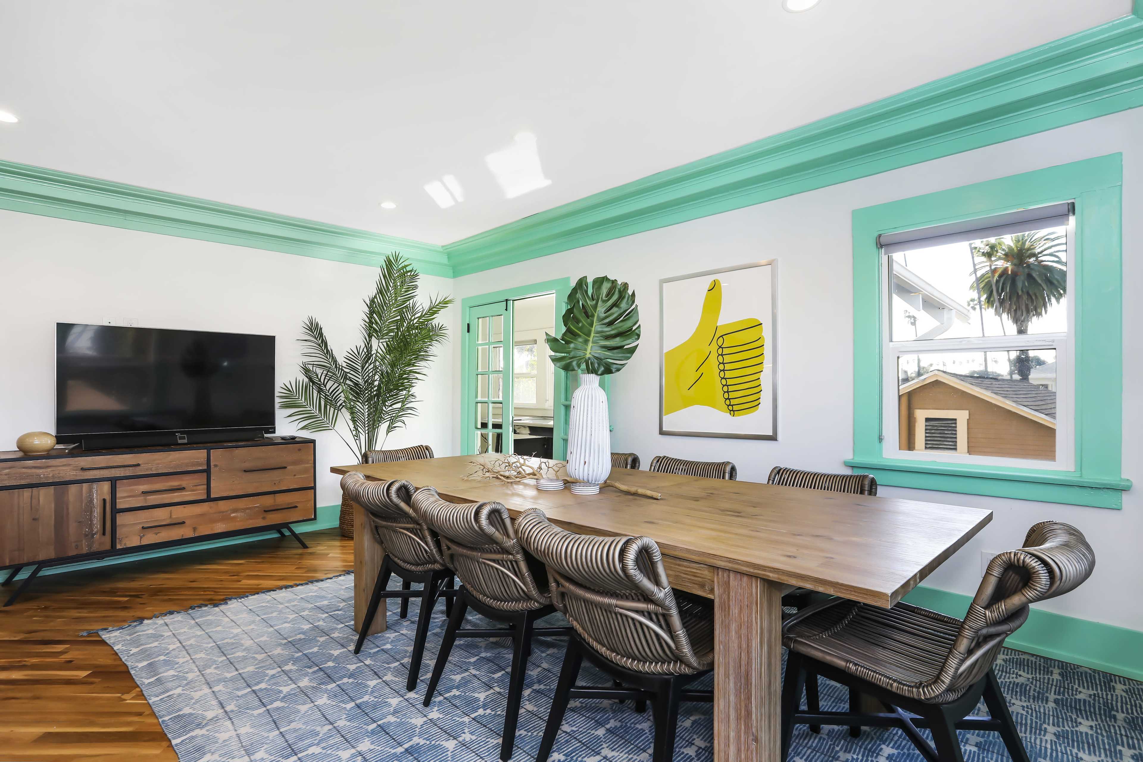 A modern dining area features a large wooden table surrounded by black wicker chairs, with a television on a wooden cabinet and bright green accents on the walls and trim.