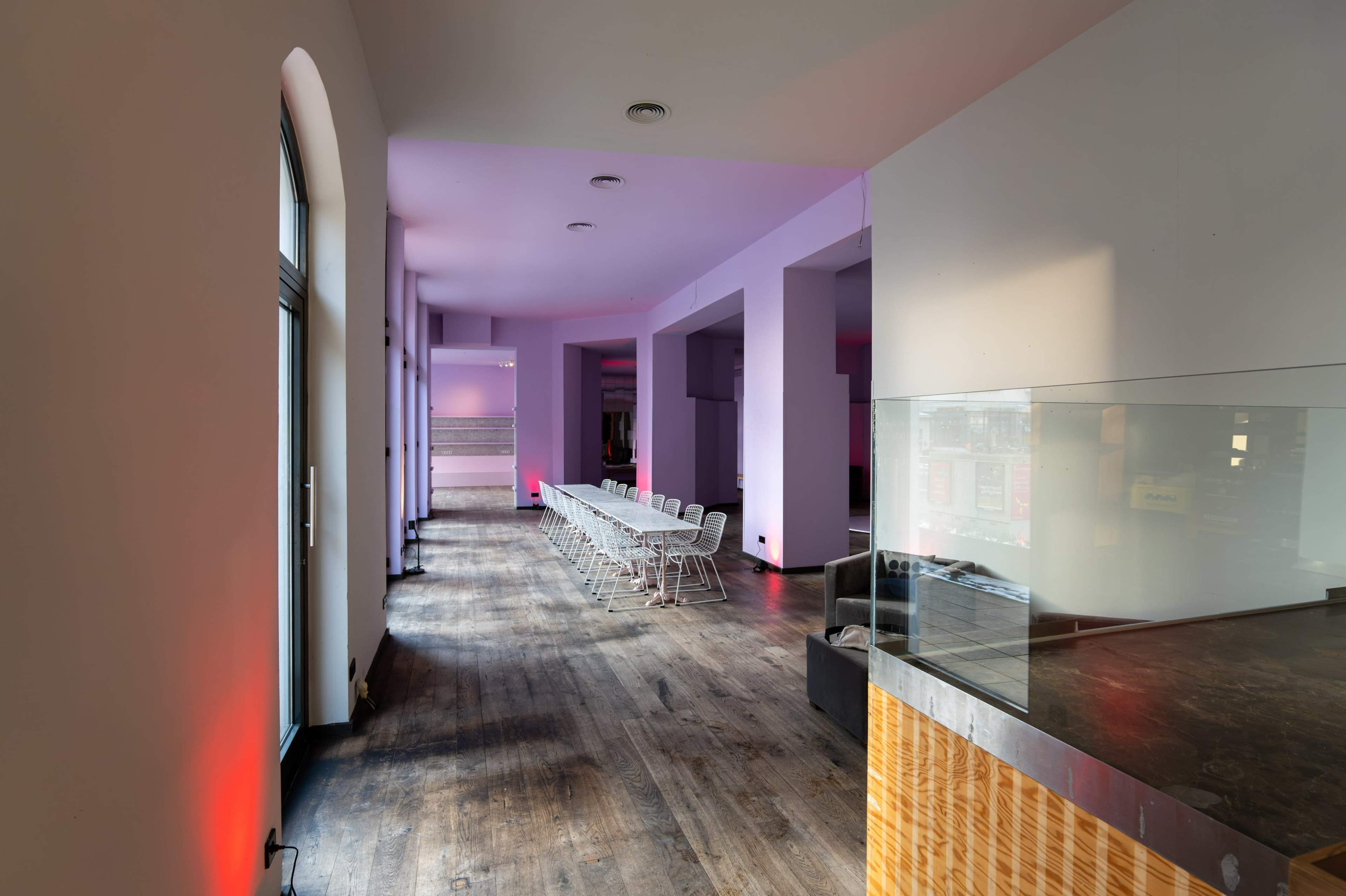 A spacious, minimalist interior with a long table lined with chairs, illuminated by soft purple lighting along the walls.