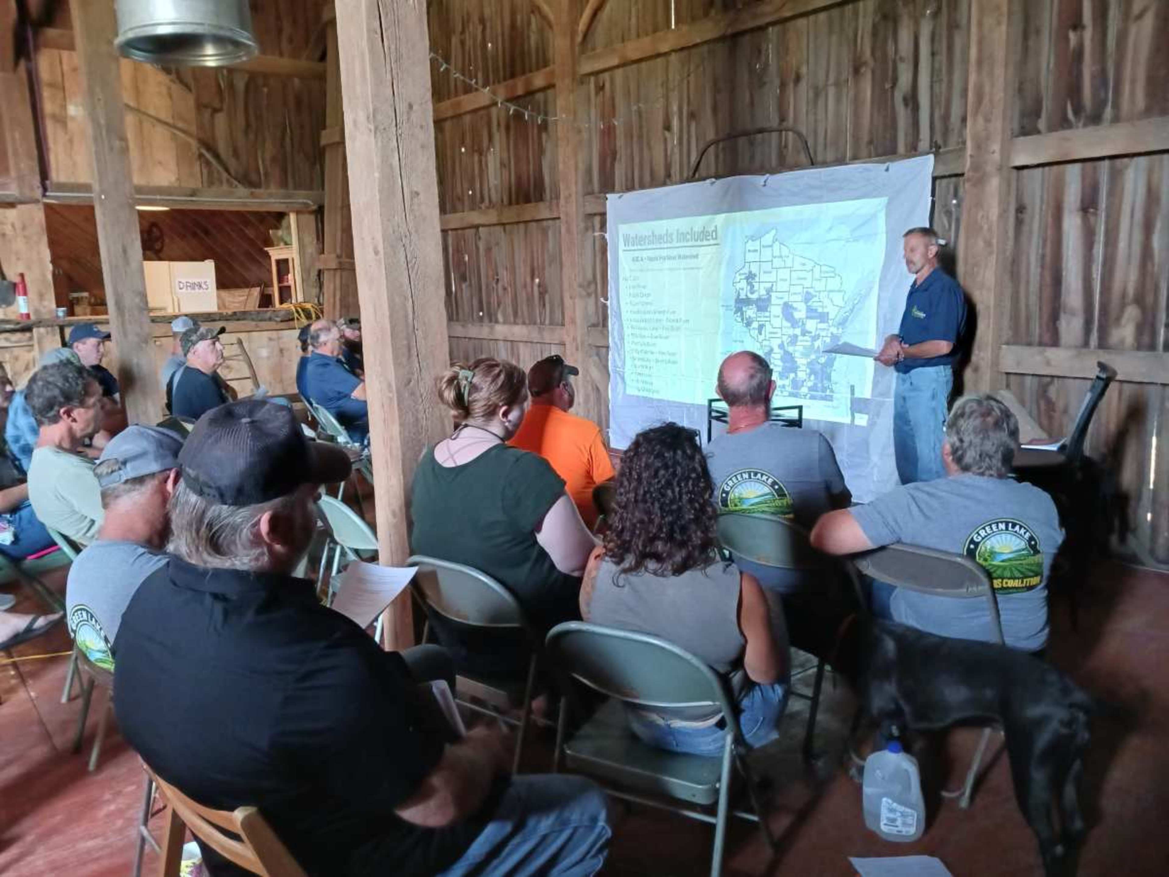 A speaker presents information to an audience seated in a rustic barn, with a map displayed in the background.