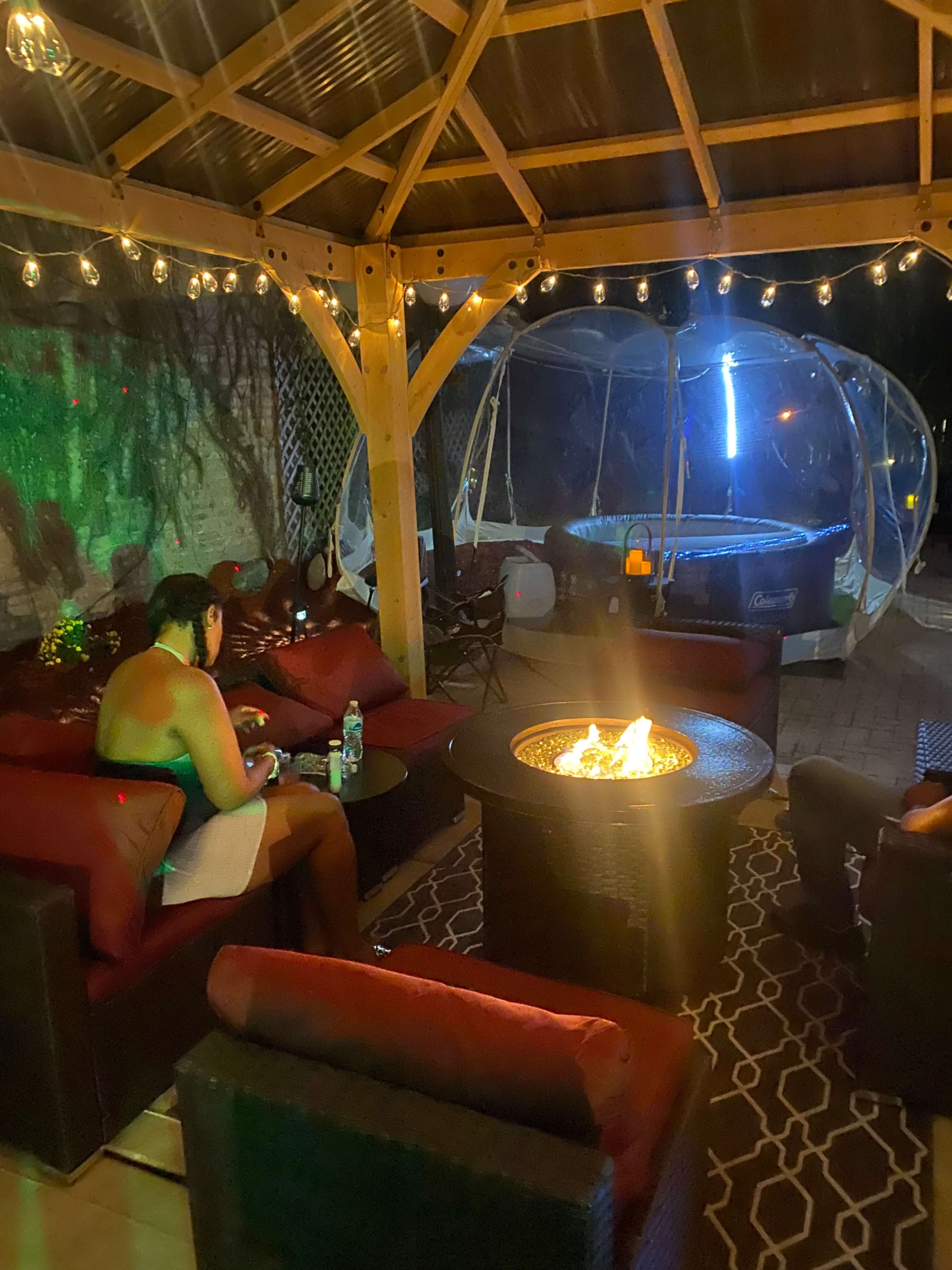 A cozy outdoor seating area with a fire pit surrounded by dark wicker furniture and string lights, adjacent to a large inflatable hot tub.