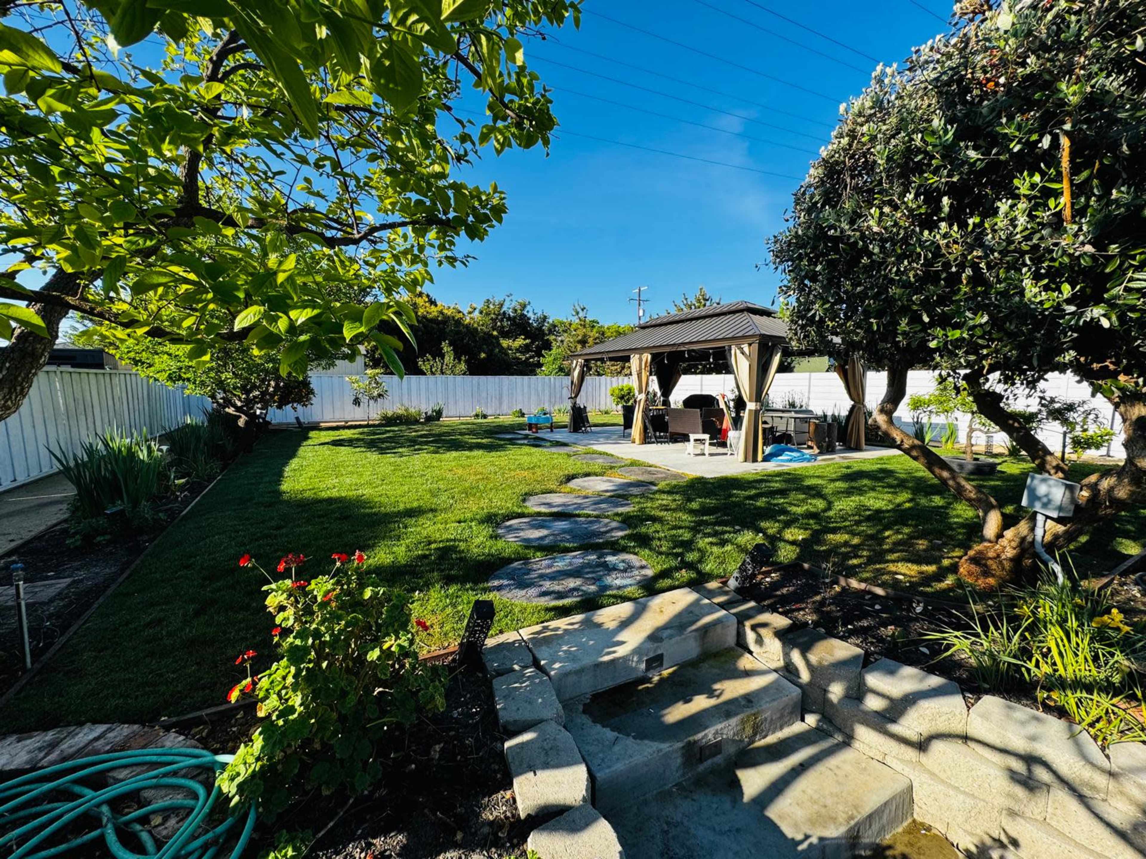 The image shows a well-manicured backyard with a patio under a pergola, surrounded by greenery and a pathway made of stones.