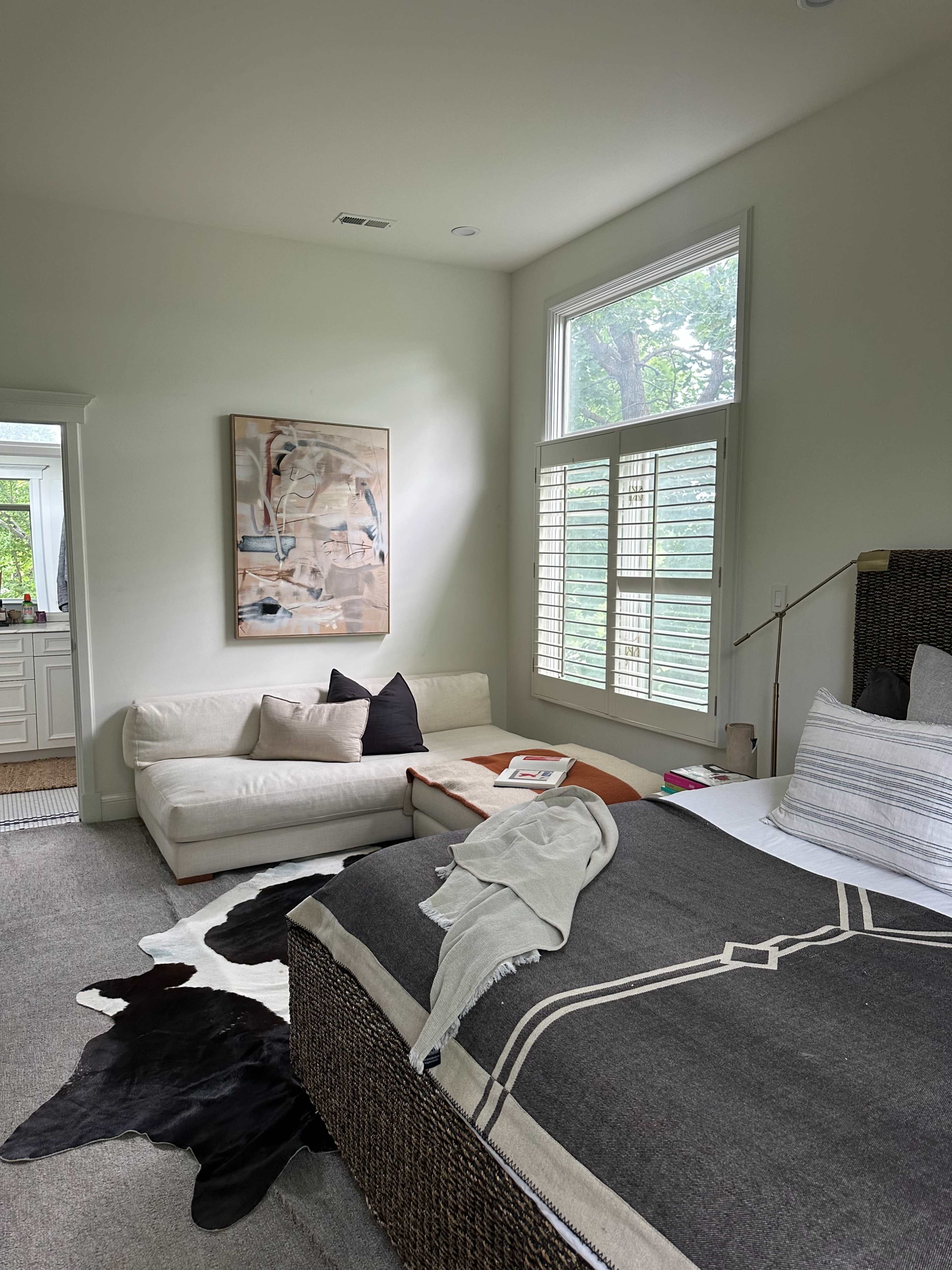 A modern bedroom features a bed with a dark throw blanket, a leather couch, and a large window with shutters, showcasing a minimalist design.