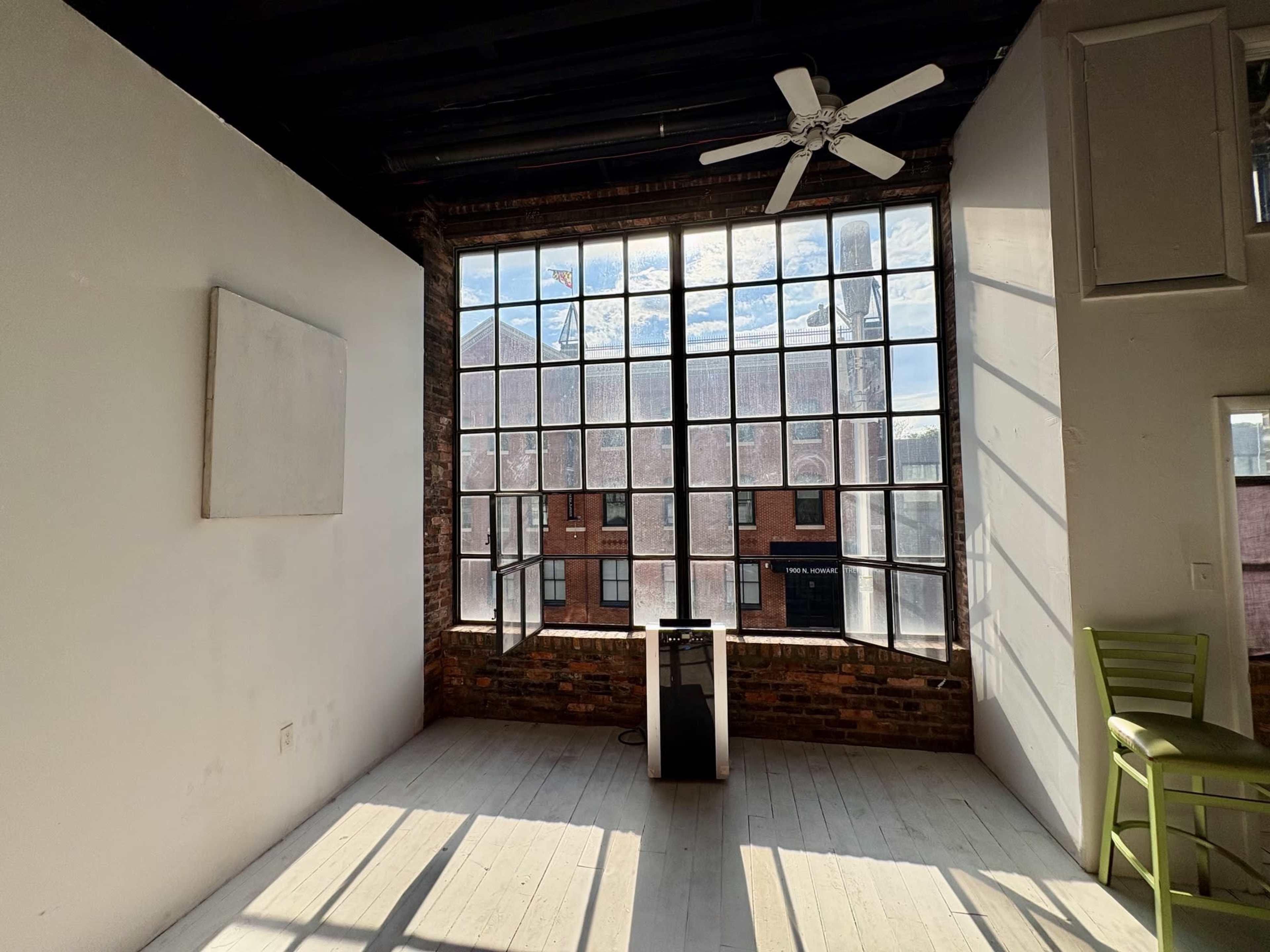 The image shows a bright, spacious room with a large industrial-style window, wooden floors, and a small speaker or device near the center.