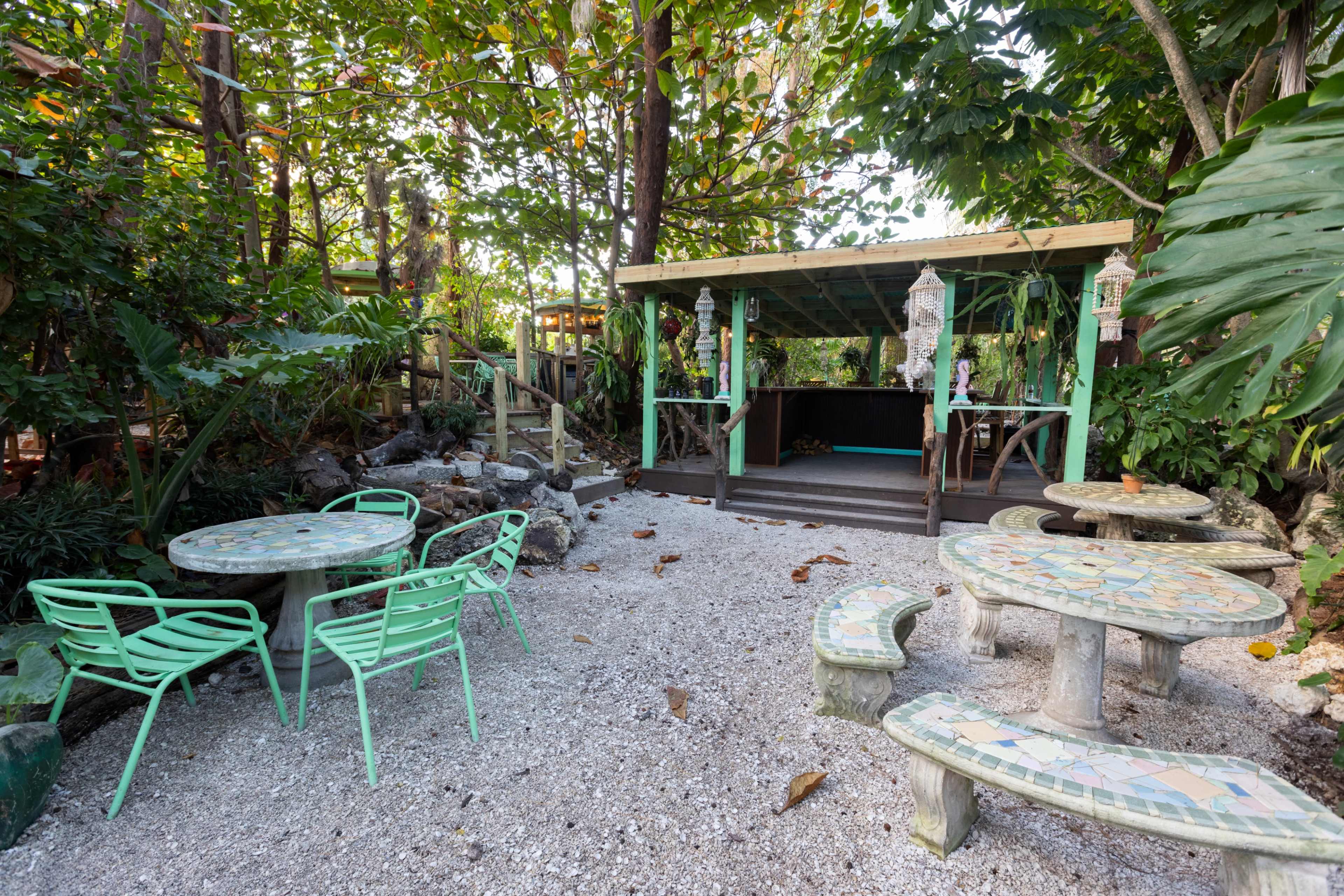 The image shows an outdoor seating area featuring mosaic-topped tables and bright green chairs, surrounded by lush greenery and a wooden structure in the background.