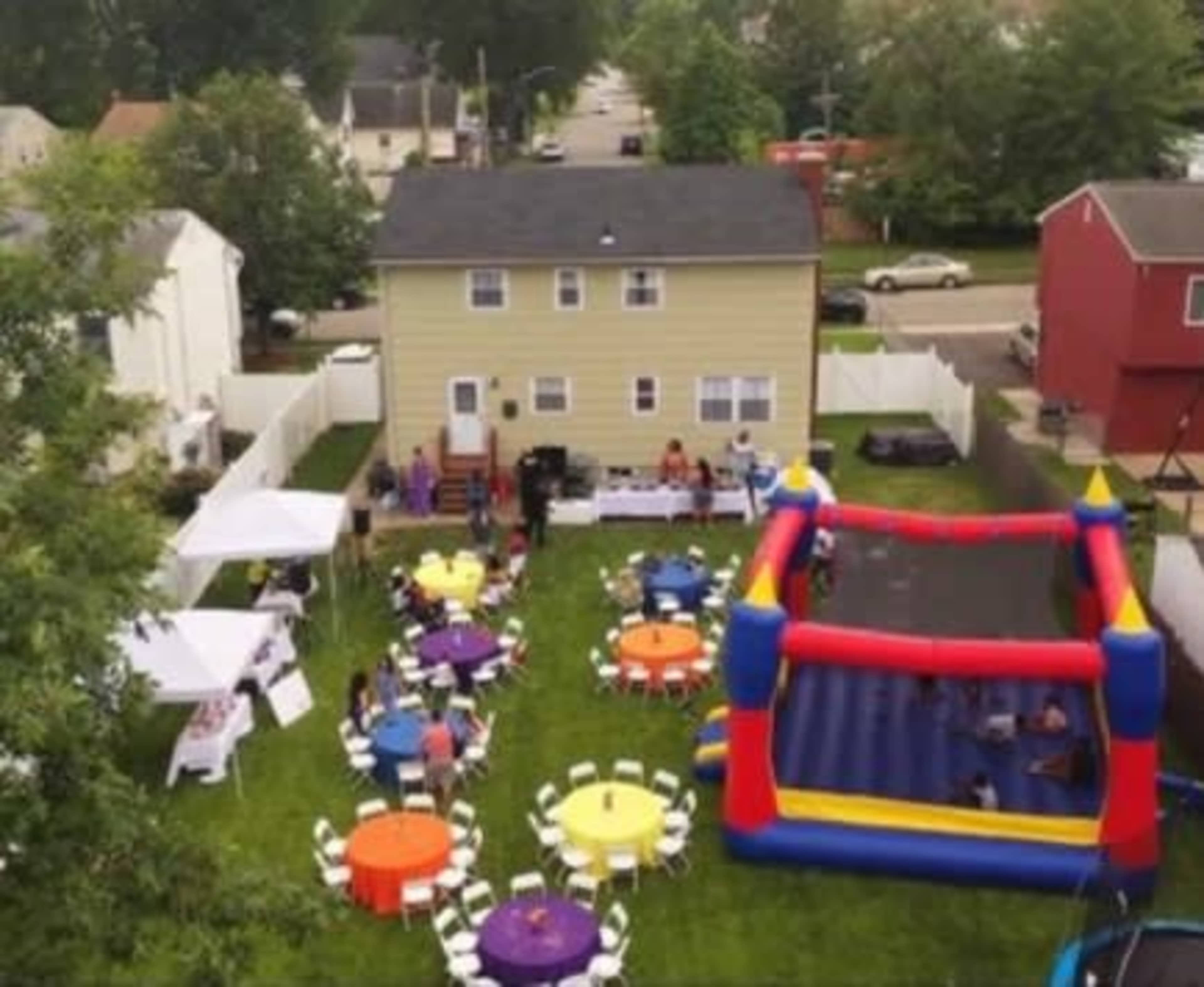 The scene shows a backyard party with colorful round tables, chairs, a bounce house, and catering tents set up for guests.