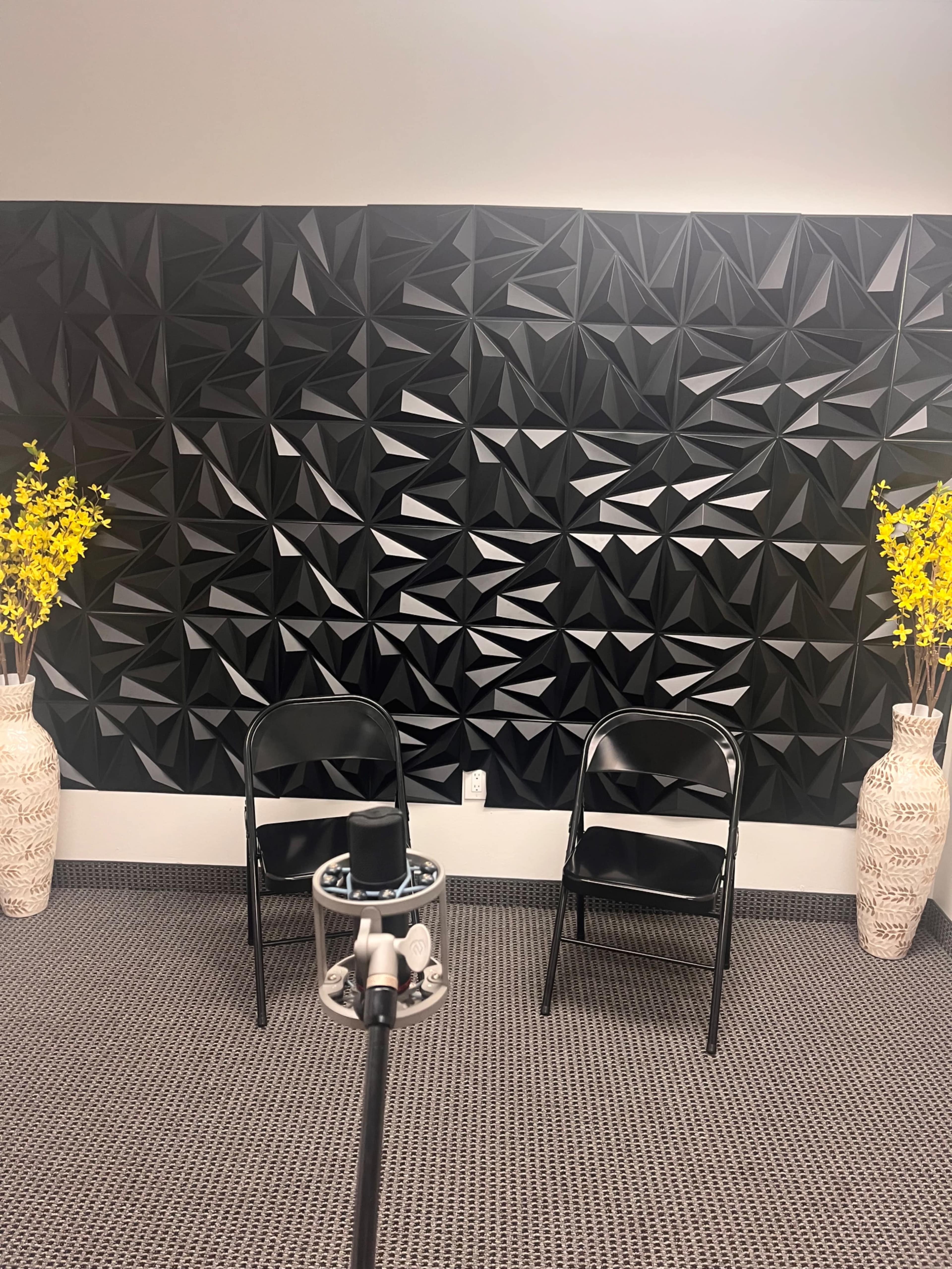 The image shows a room with a textured black wall, two black chairs, a small round table, and two decorative vases with yellow flowers.