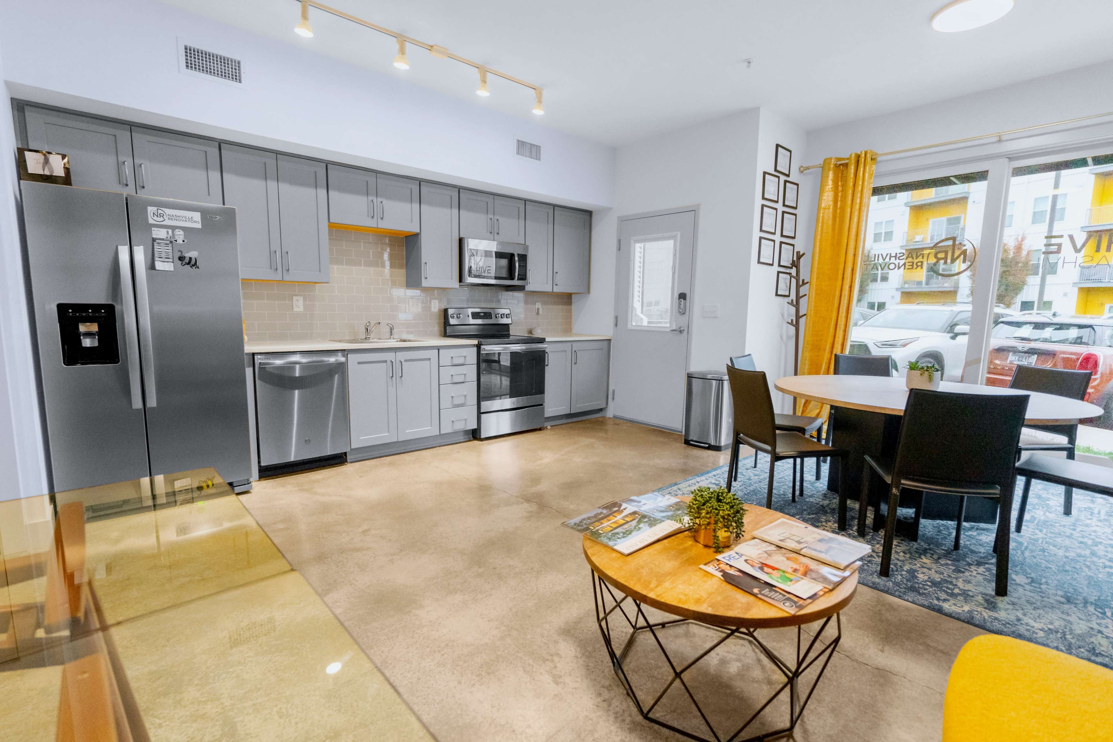 A modern kitchen and dining area featuring stainless steel appliances, a dining table, and large windows with yellow curtains.