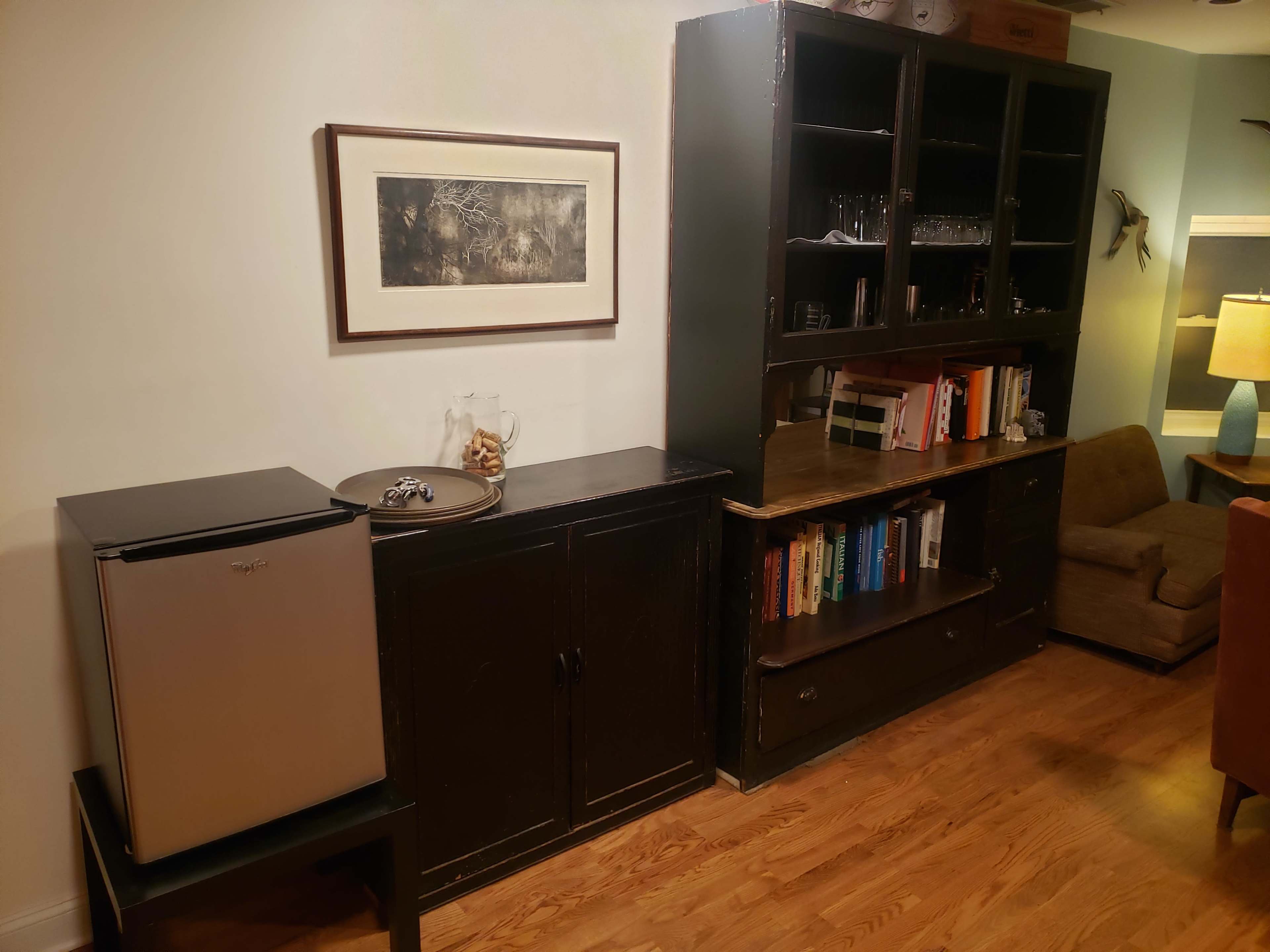 A corner of a room featuring a small fridge on a stand next to a dark wooden cabinet with shelves holding books and glassware, along with a framed picture on the wall above.