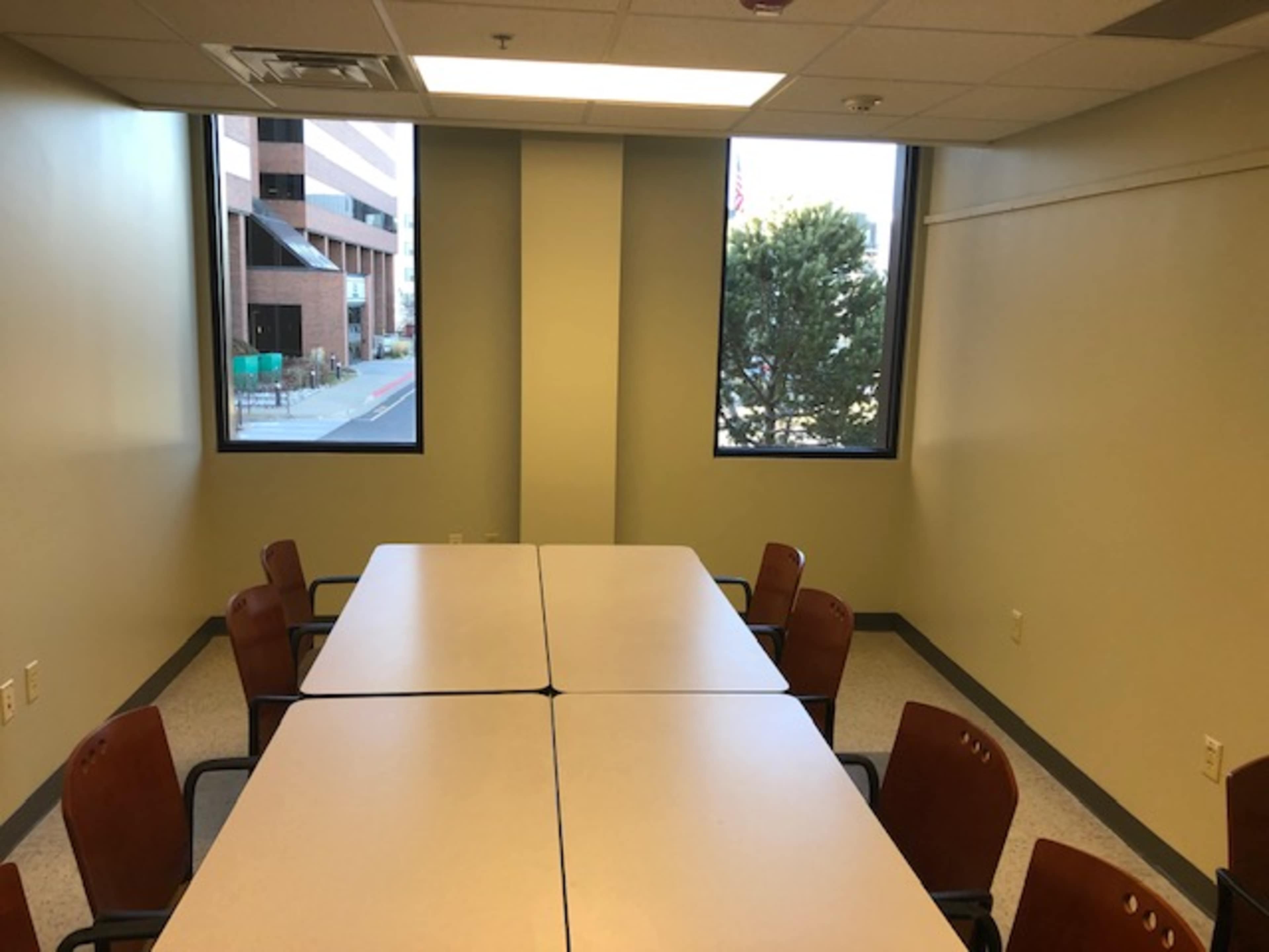 A meeting room with a long table surrounded by several chairs and large windows on two walls.