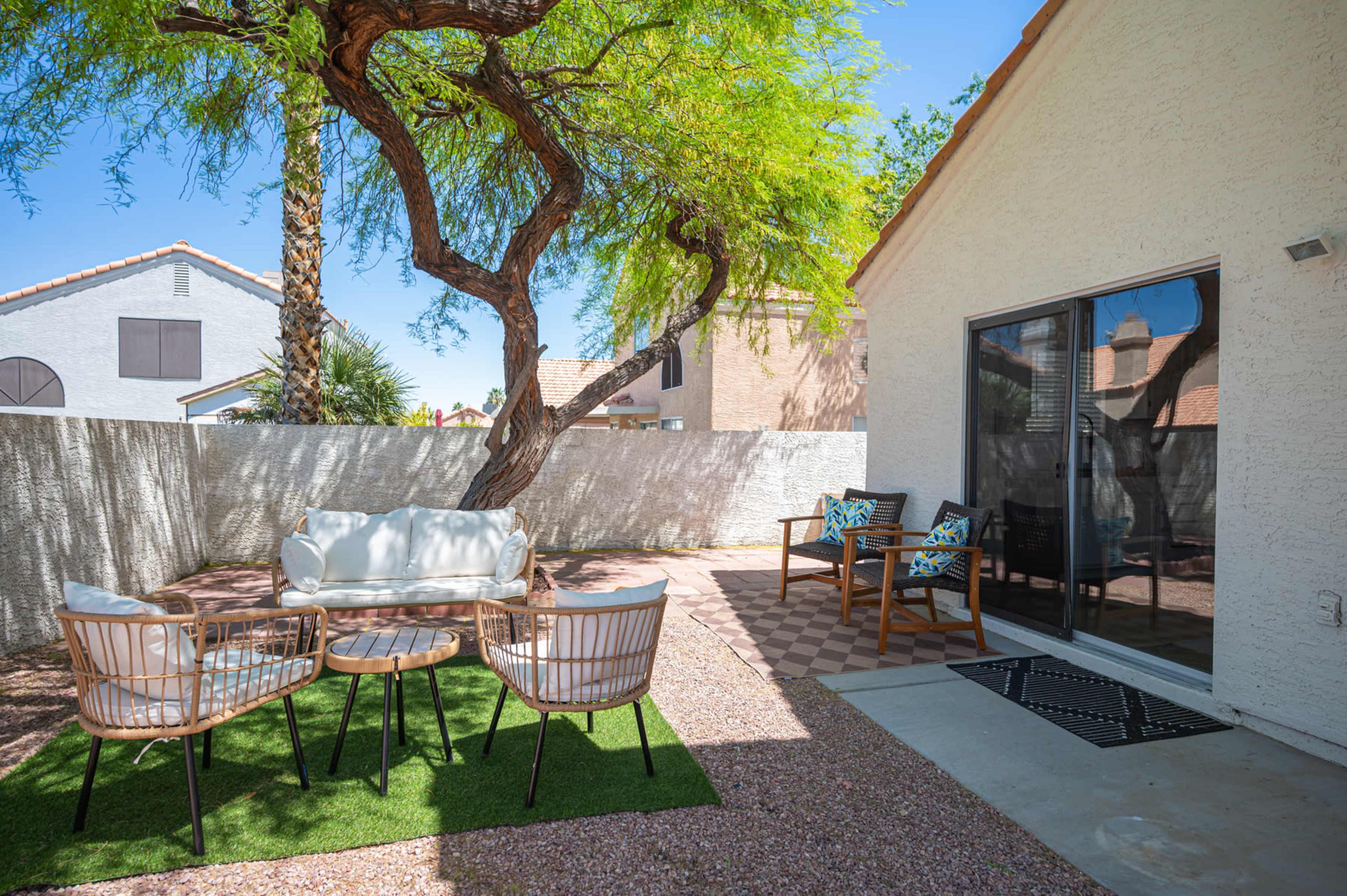 A patio area features a round table with four chairs on grass, accompanied by a couch and two side chairs against a house wall.