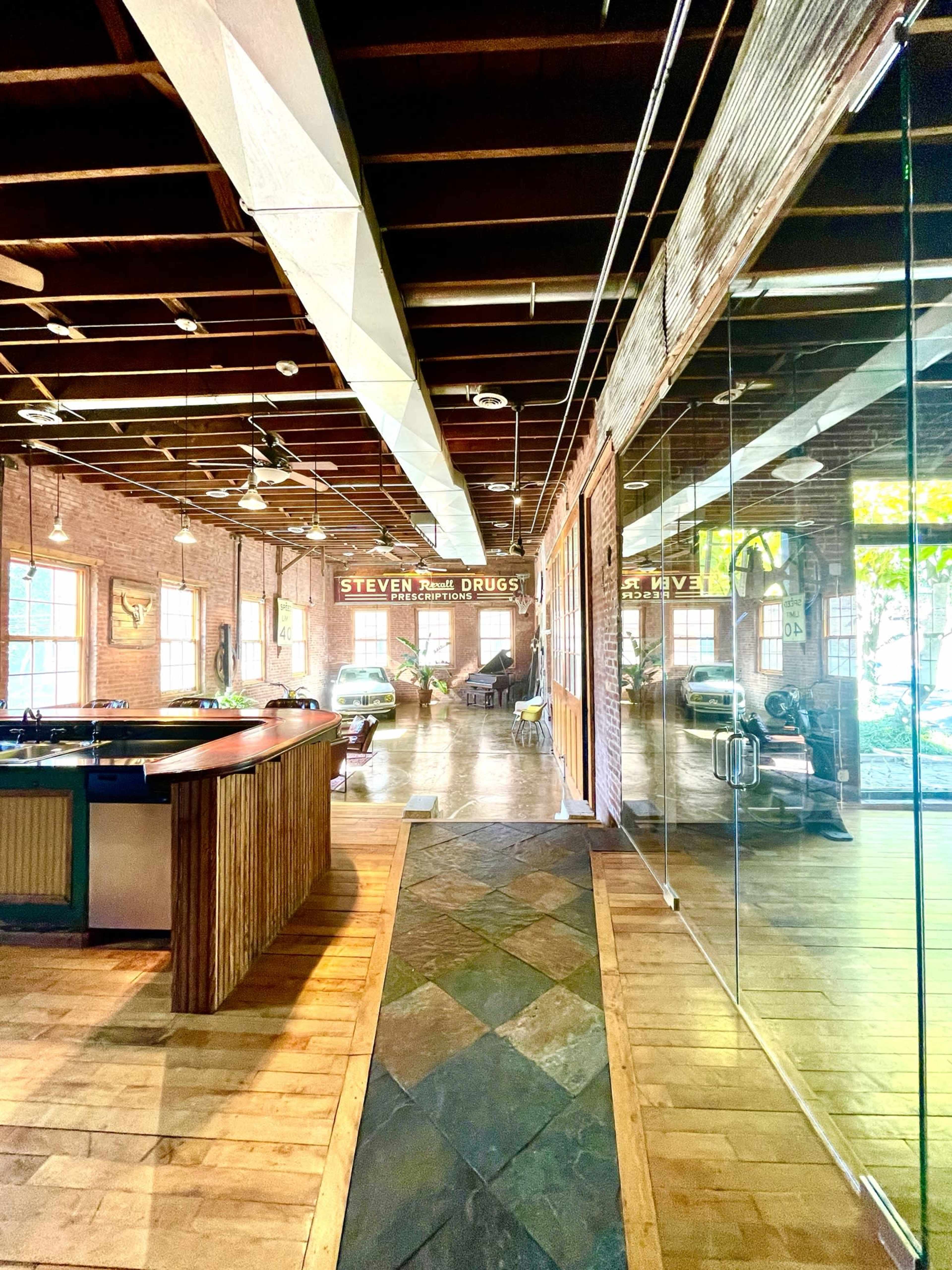 A spacious interior of a rustic co-working space with exposed wooden beams and glass walls, featuring a long communal table and seating areas.