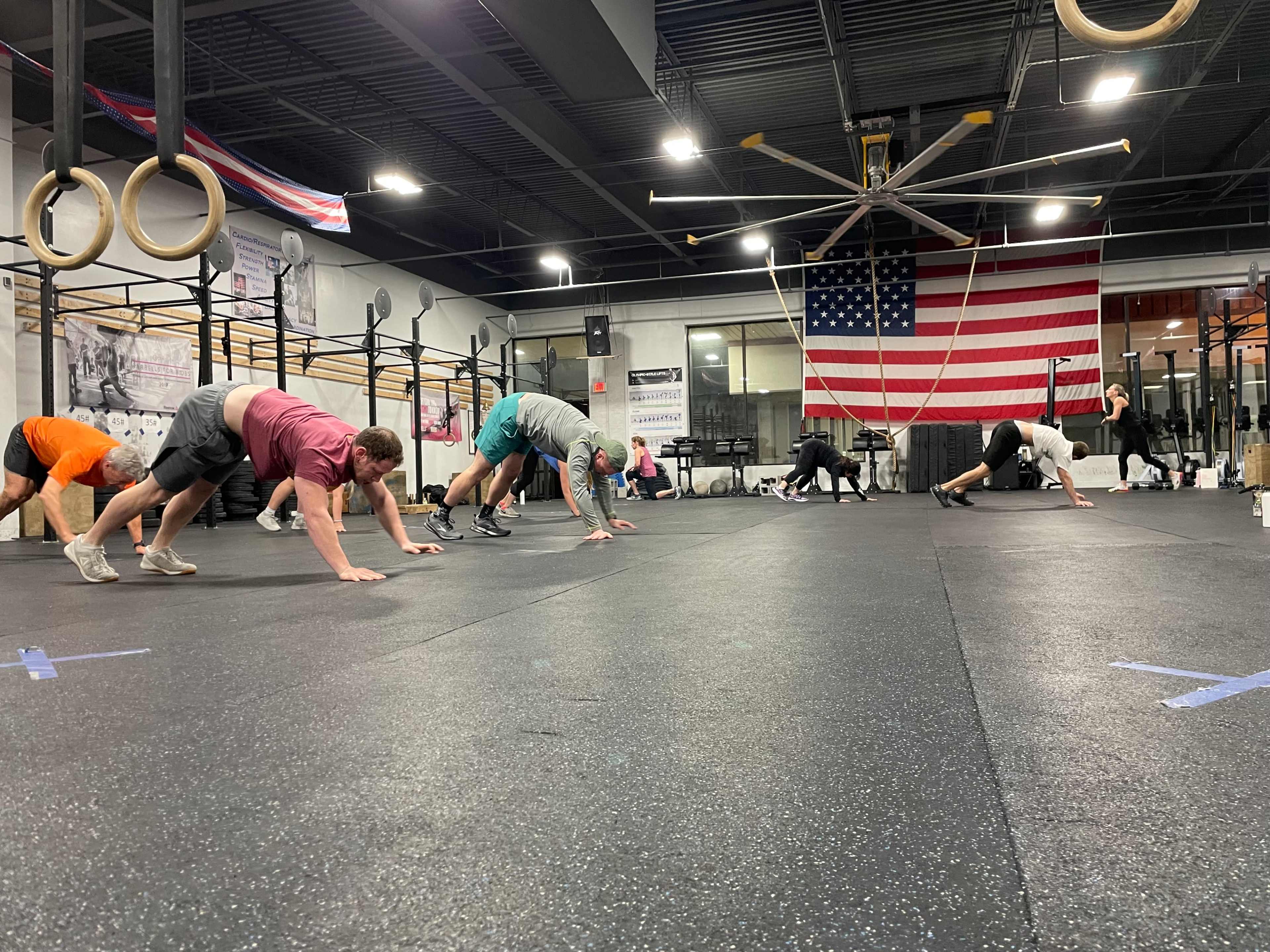 A group of individuals performs exercises on a rubberized gym floor, with an American flag hanging in the background.