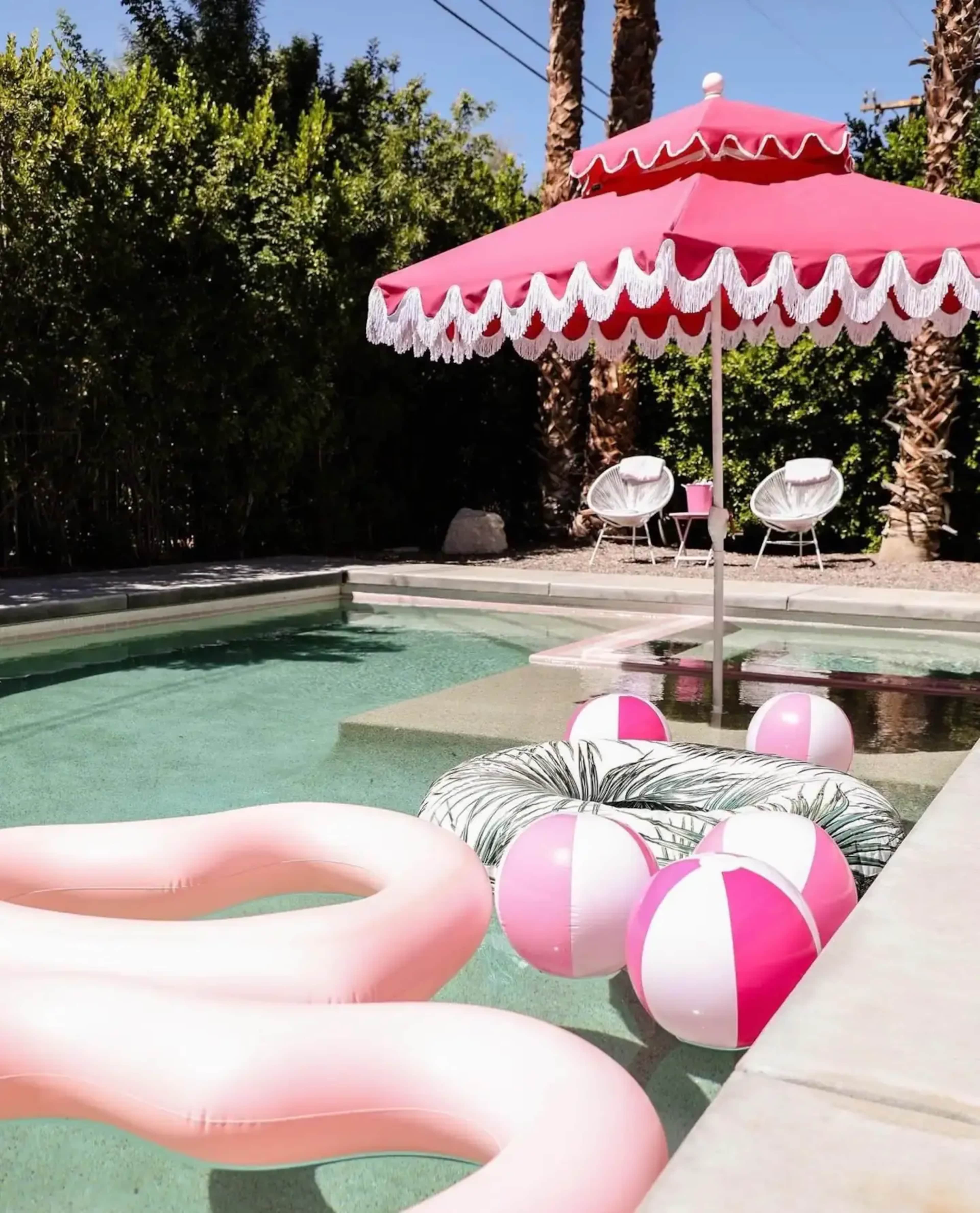 A pink parasol shades a poolside area featuring inflatable pool toys and colorful beach balls in the water.