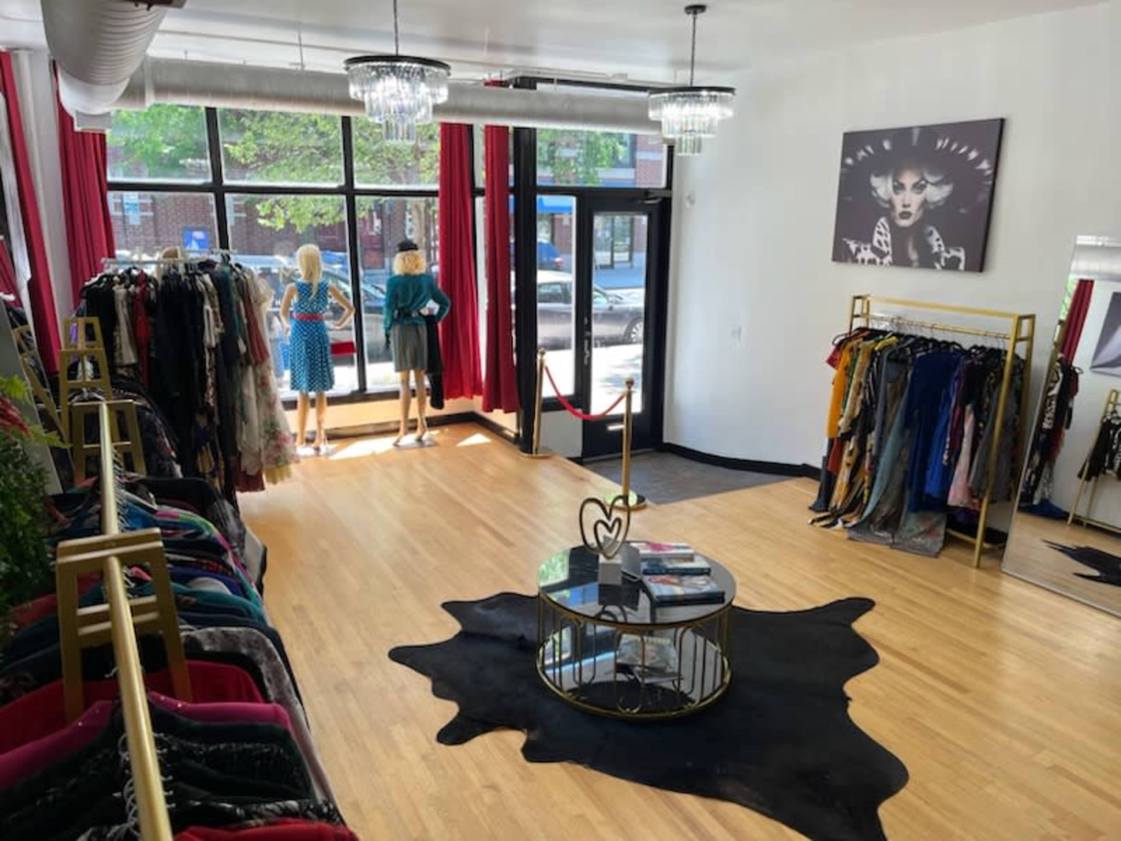 The image shows a modern boutique with clothing racks, a central coffee table, and mannequins dressed in outfits, against a backdrop of large windows and red curtains.