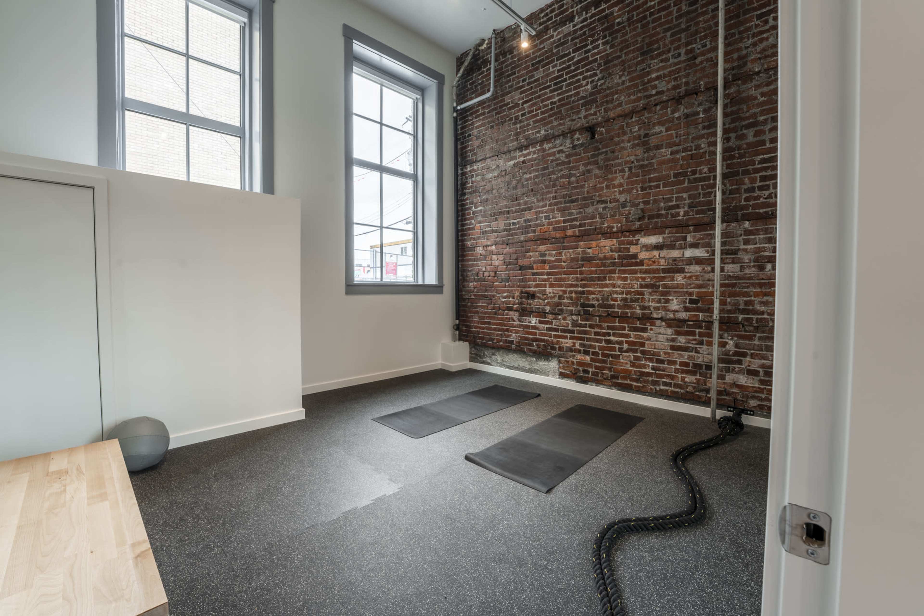 A minimalistic room with two exercise mats on a black floor, a brick wall, and large windows.