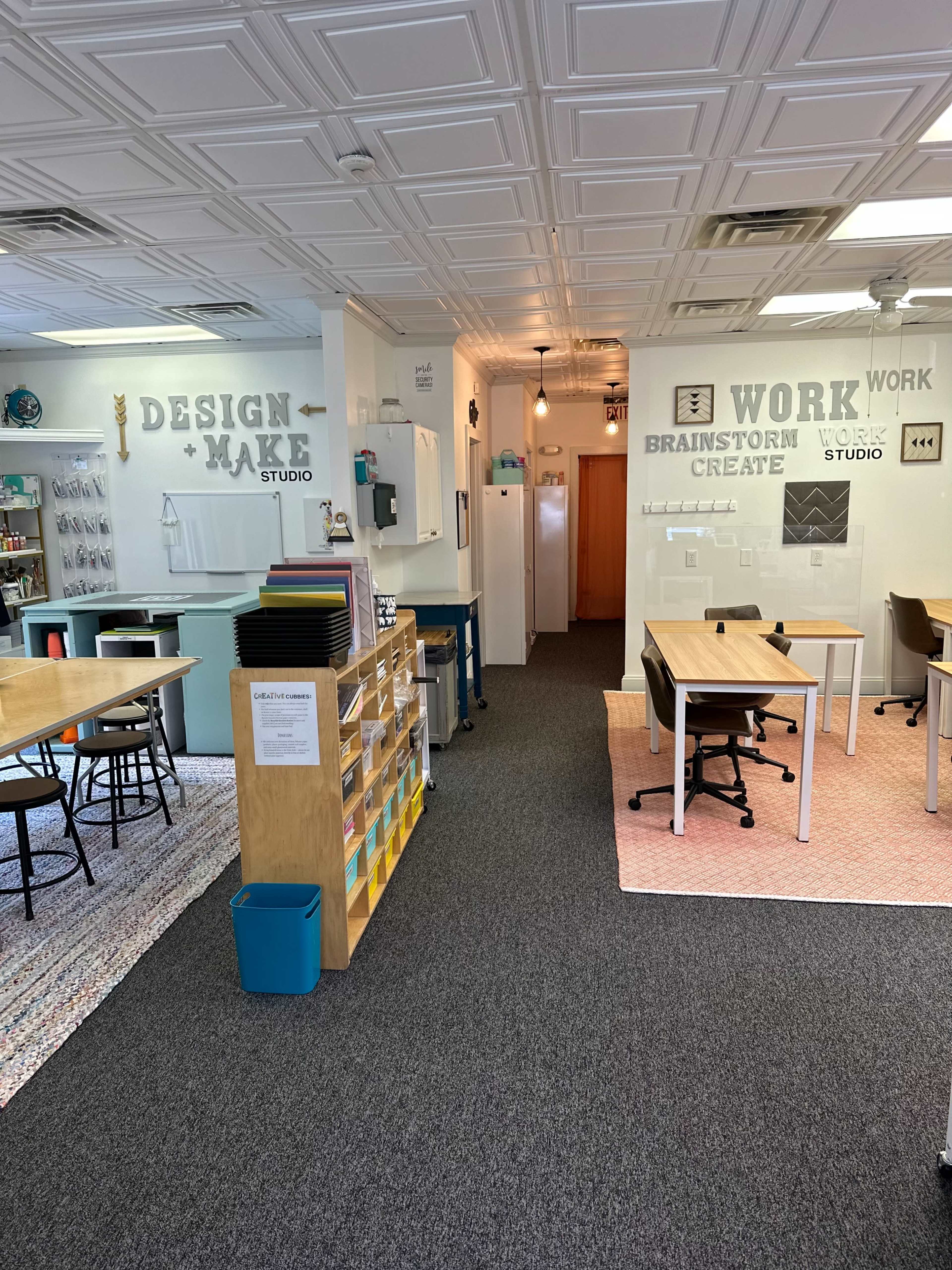 A spacious classroom or studio area featuring organized workstations, a storage unit with colorful boxes, and walls labeled with words like "DESIGN," "MAKE," "WORK," and "CREATE."