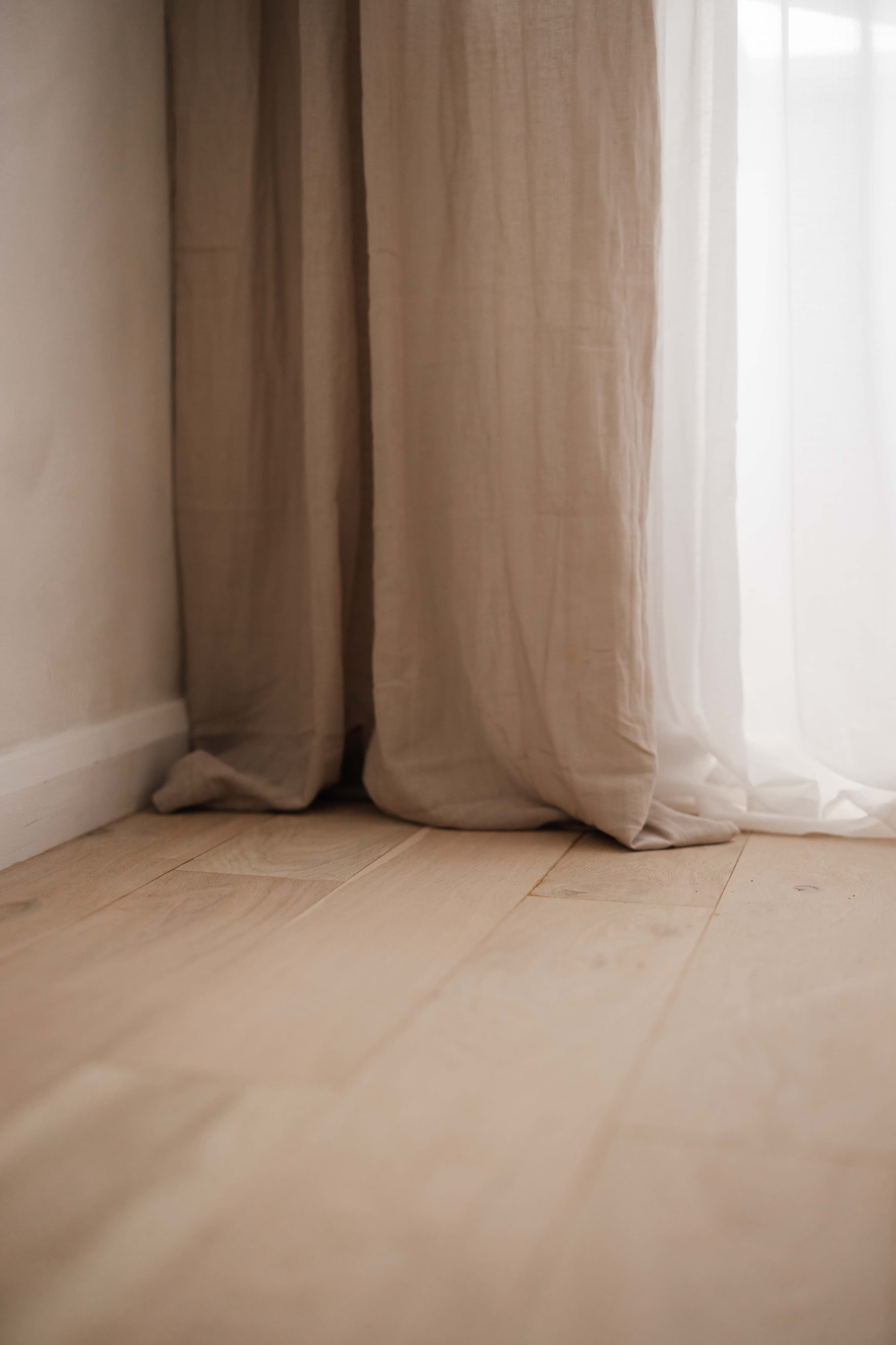 The image shows a corner of a room with light-colored wooden flooring and sheer curtains draping beside a wall.