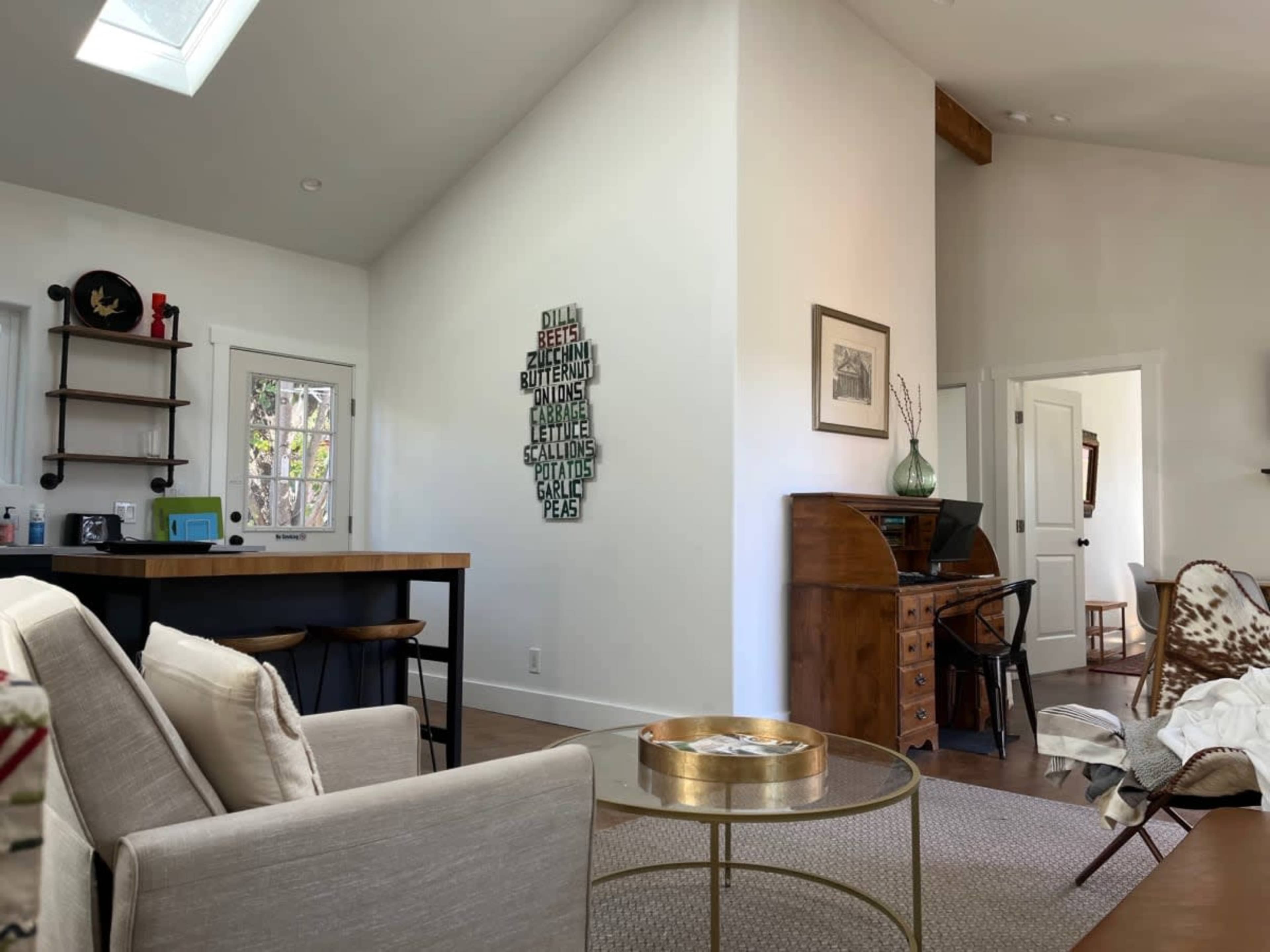 A modern living space features a partially open layout with a kitchen, a dining area, and a workspace, characterized by white walls and wooden accents.