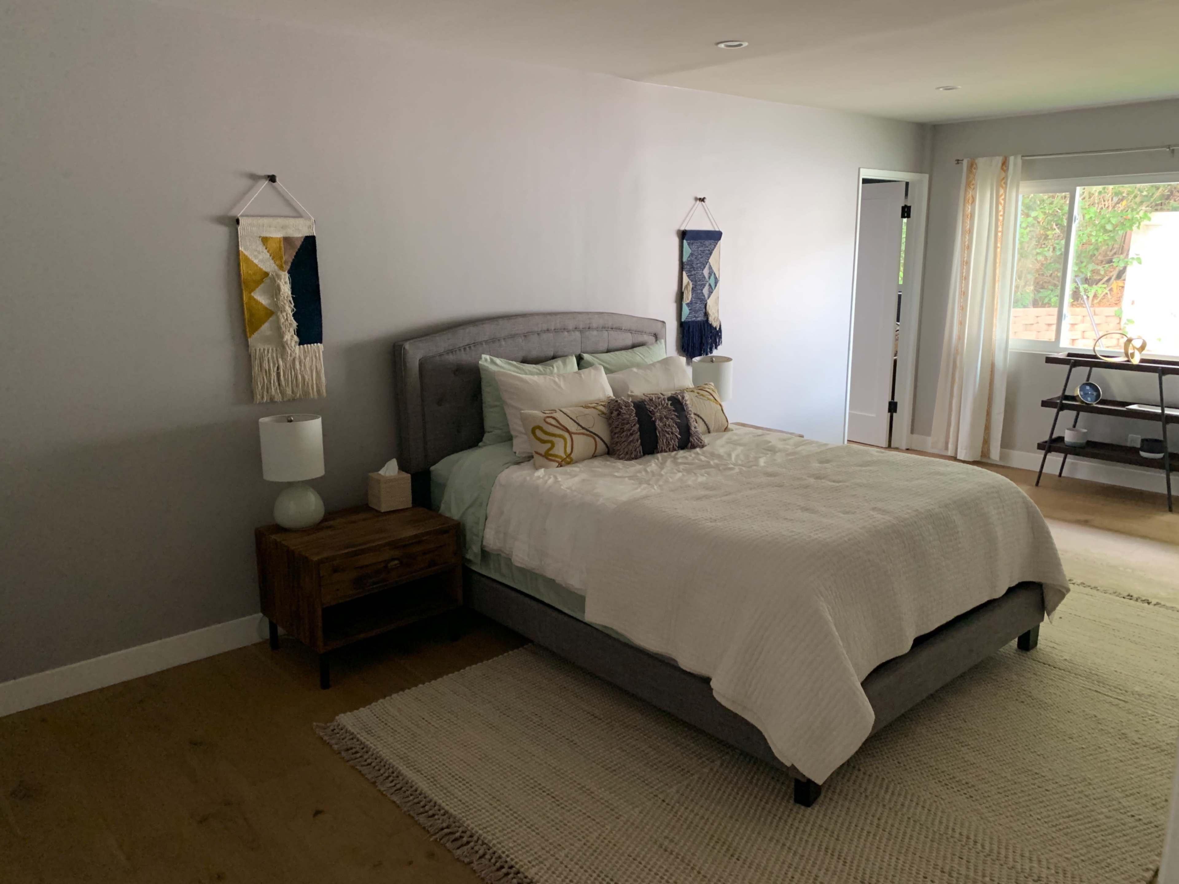A neatly arranged bedroom features a gray upholstered bed, a wooden nightstand, and decorative wall hangings beside a window.