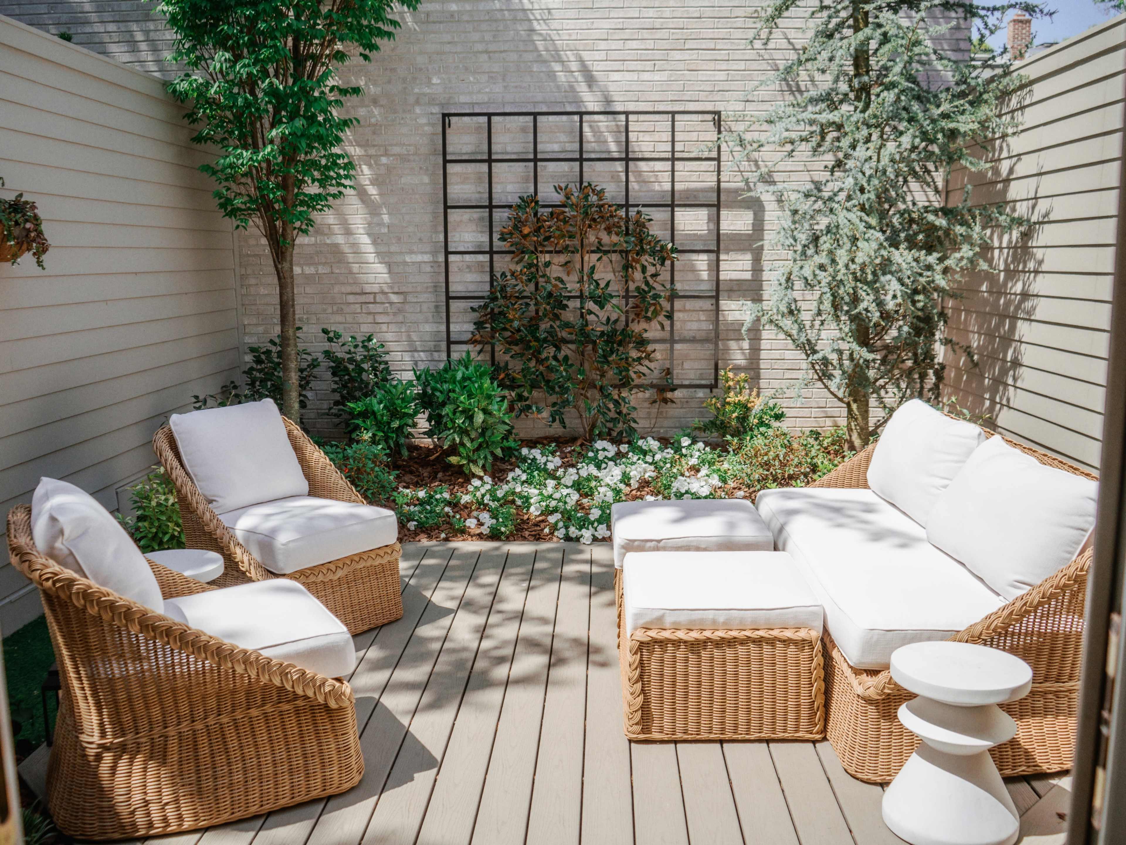 A small outdoor seating area with wicker chairs and a sectional sofa, surrounded by lush greenery and flowers.