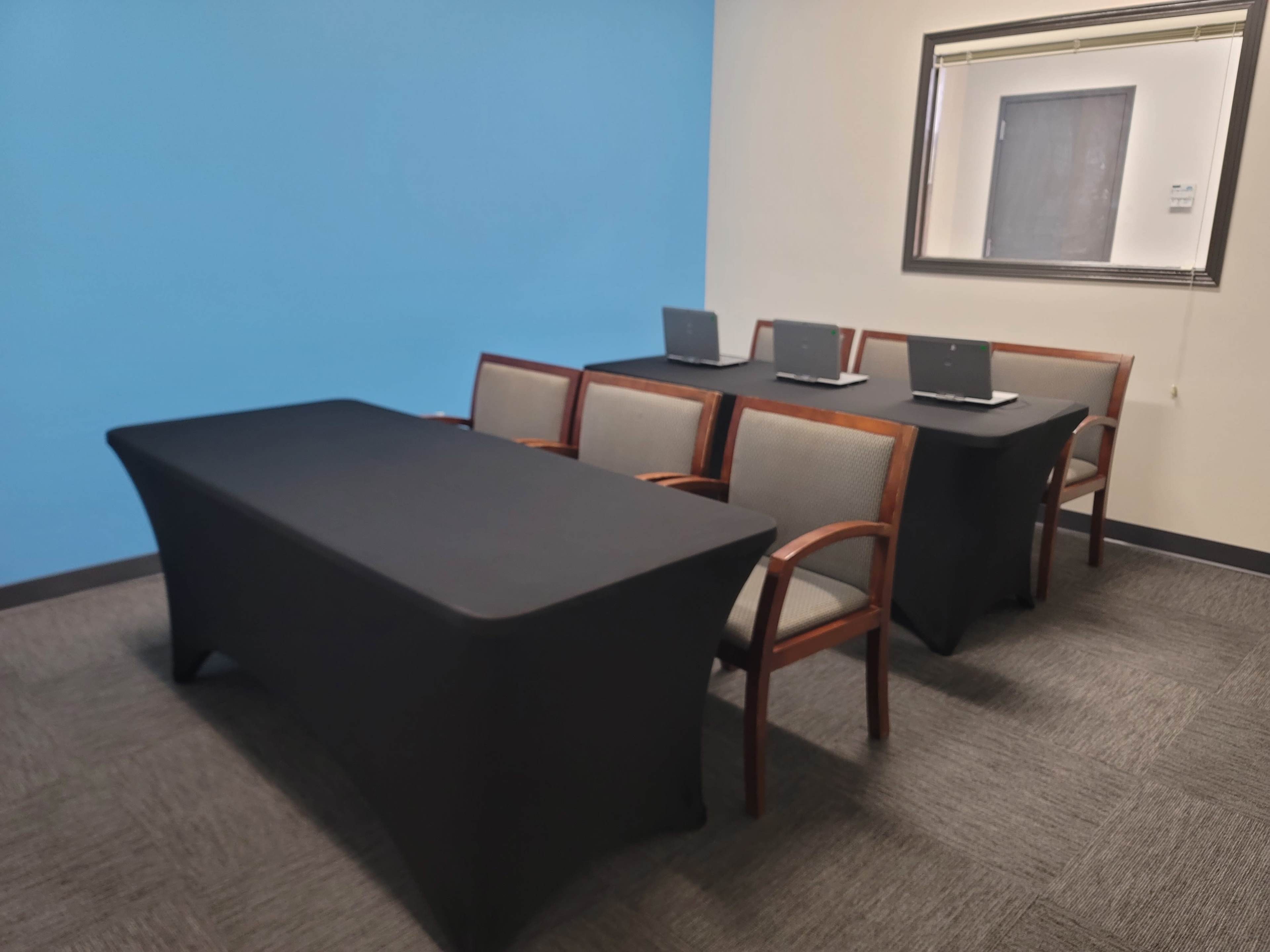 A meeting room features a long black table at the front and five chairs with laptops arranged neatly behind it against a blue wall.