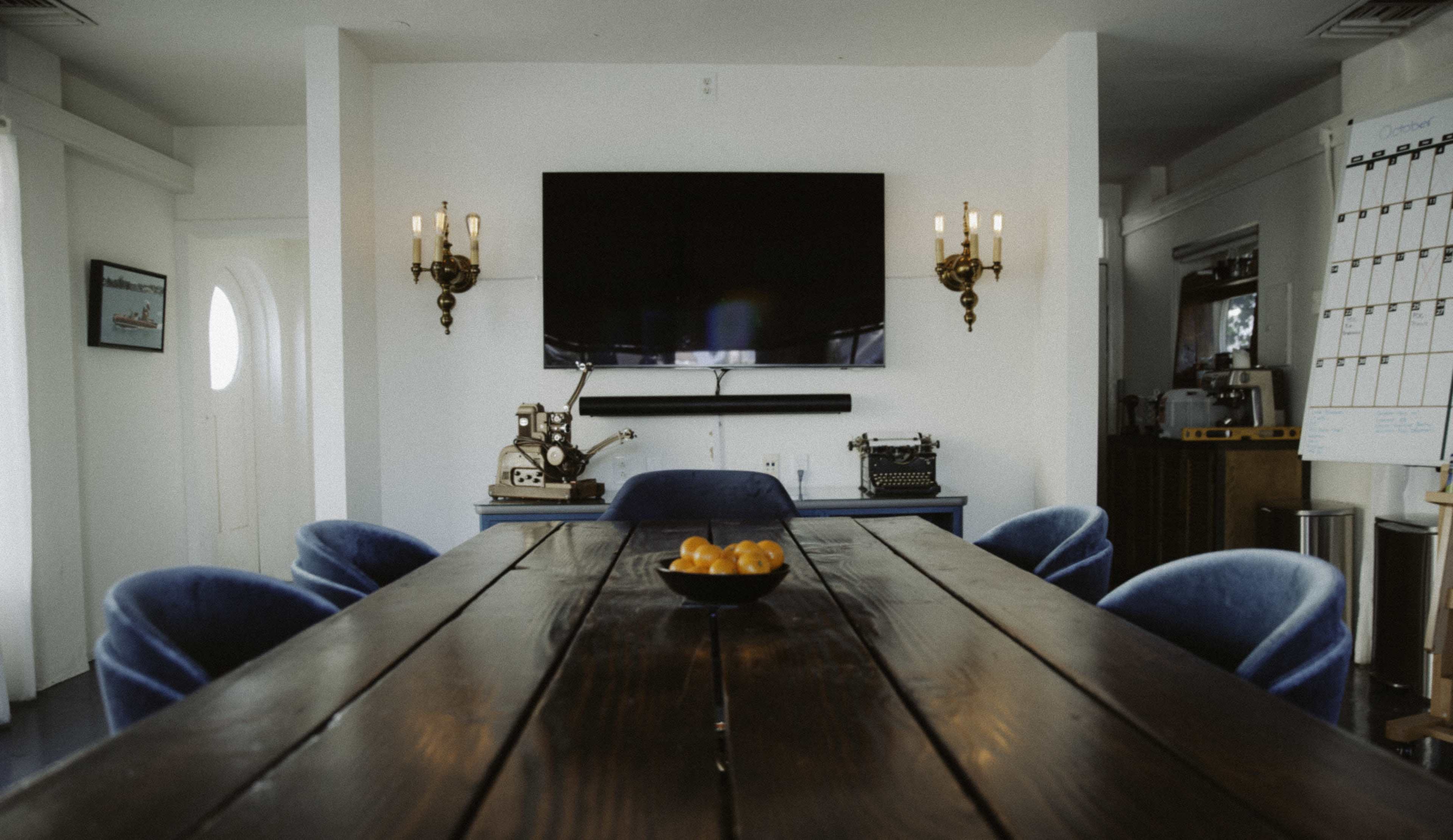 The image shows a modern dining room with a long wooden table, blue upholstered chairs, a large flat-screen TV mounted on the wall, and some decorative pieces.