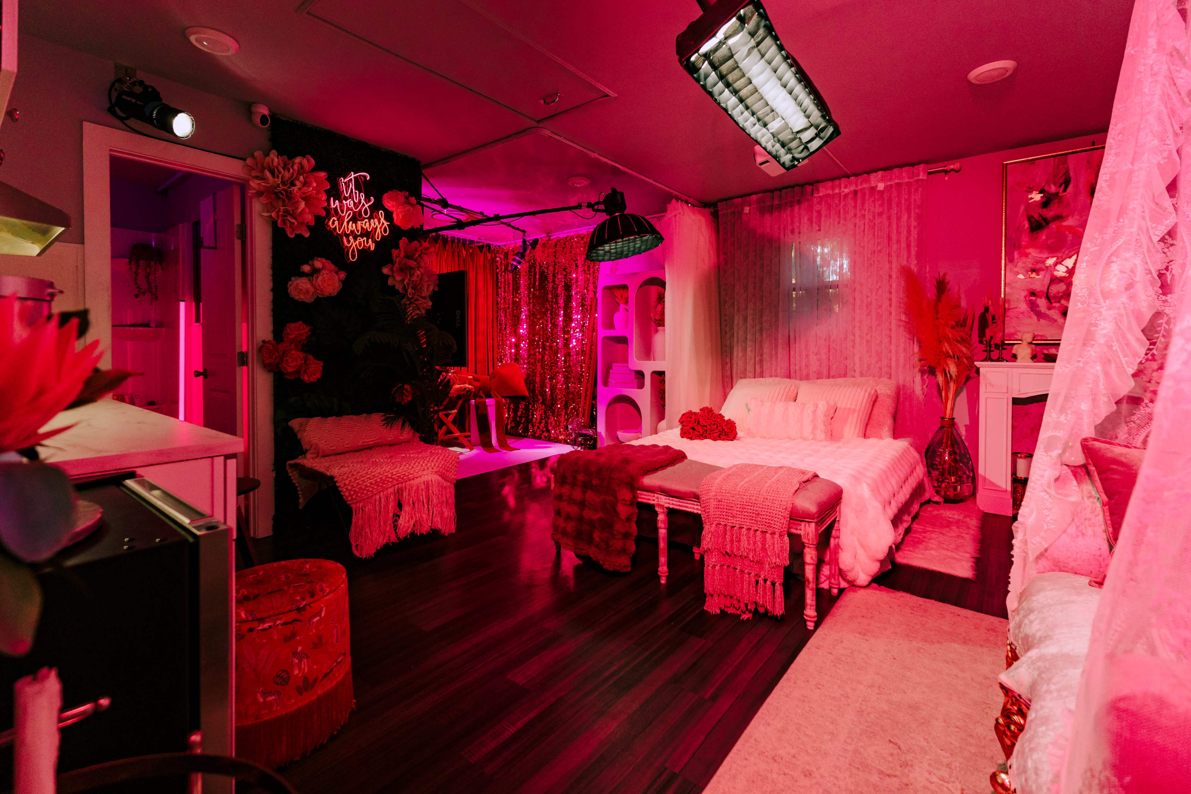 The image depicts a vibrant, pink-lit room featuring a bed with decorative pillows, a seating area, and various stylish furnishings against a backdrop of sequined curtains.