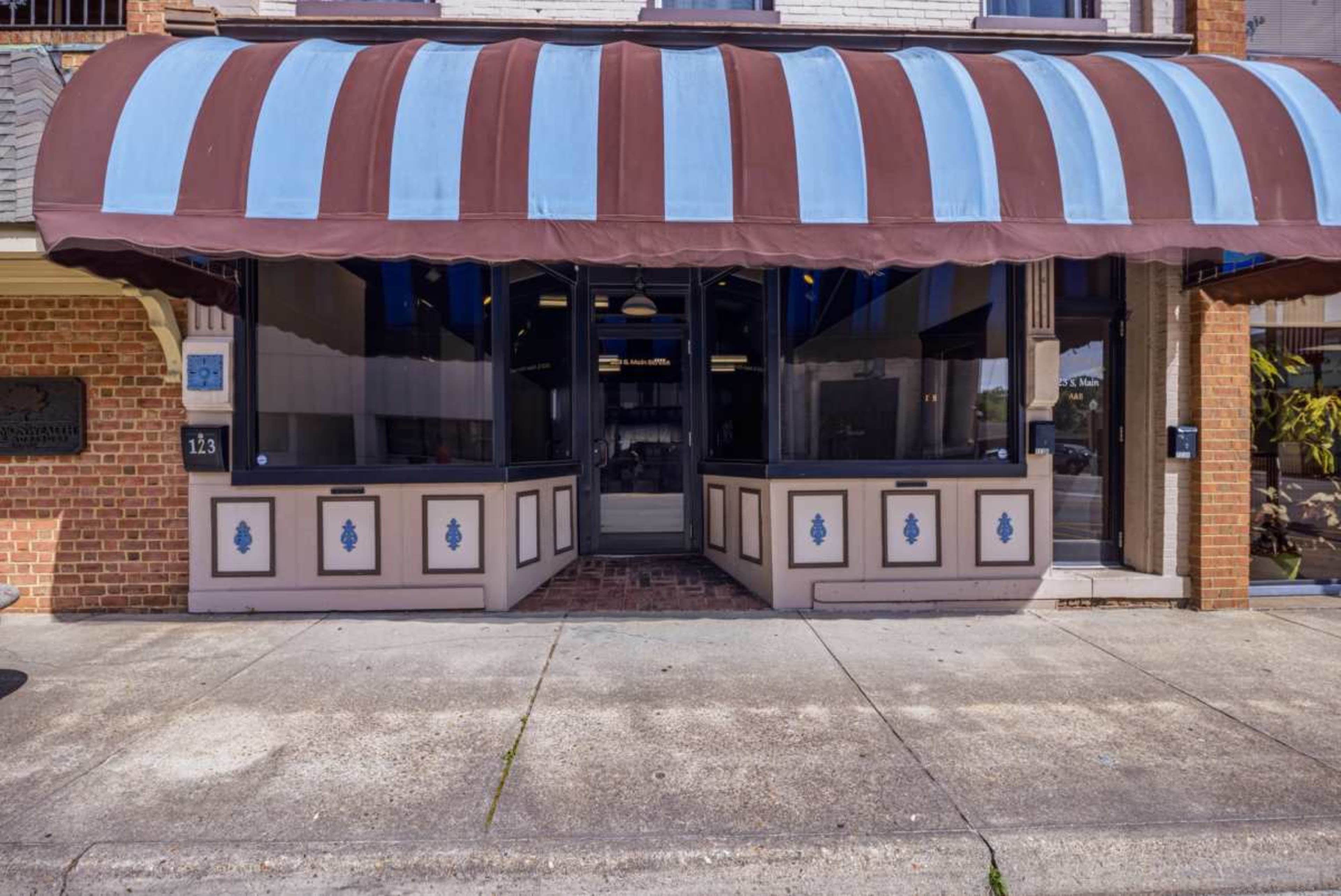 The image shows a storefront with a brown and blue striped awning, featuring a large glass window and a brick sidewalk in front.