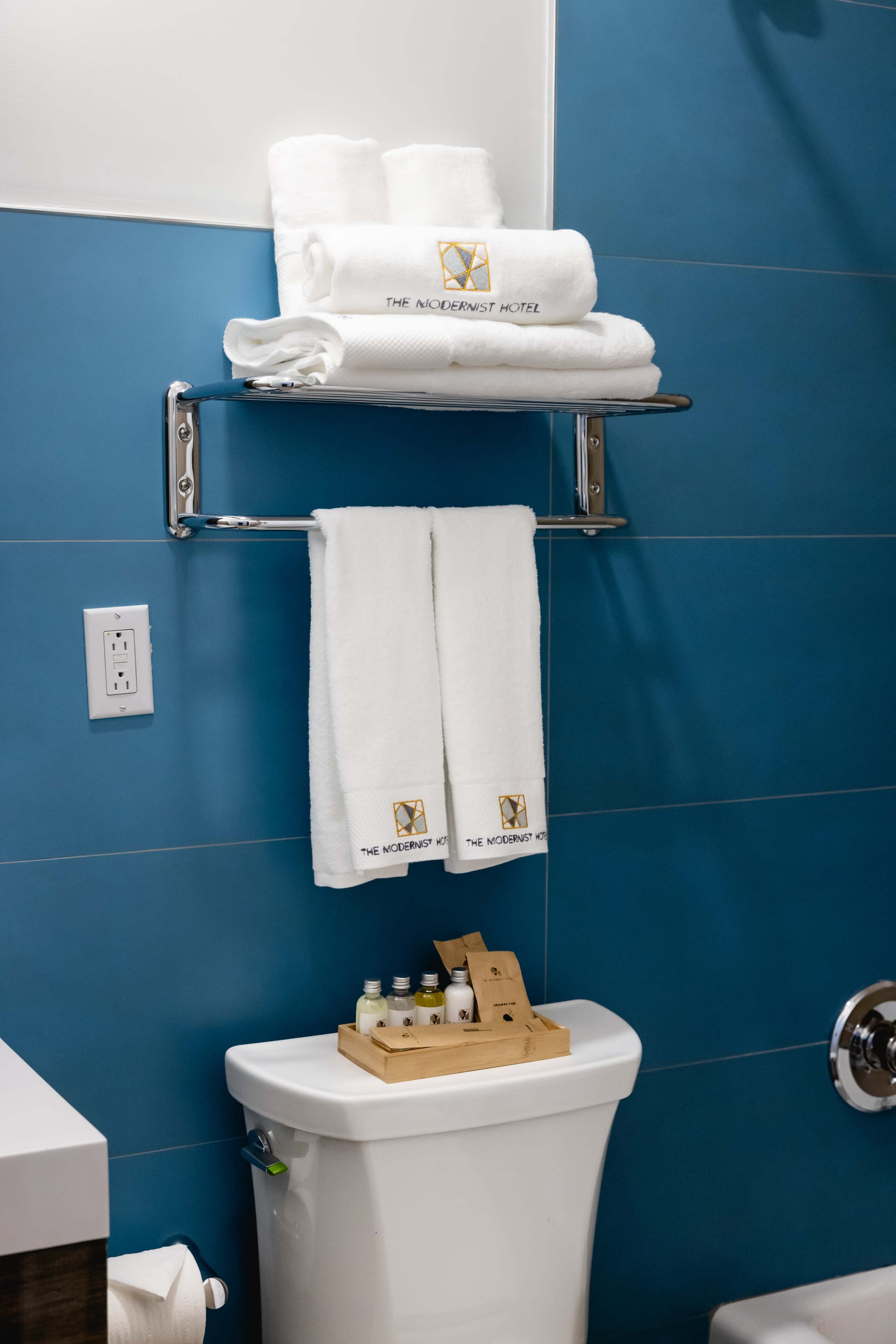 A neatly arranged bathroom displays white towels on a chrome rack above a toilet, accompanied by a wooden tray of miniature toiletries.
