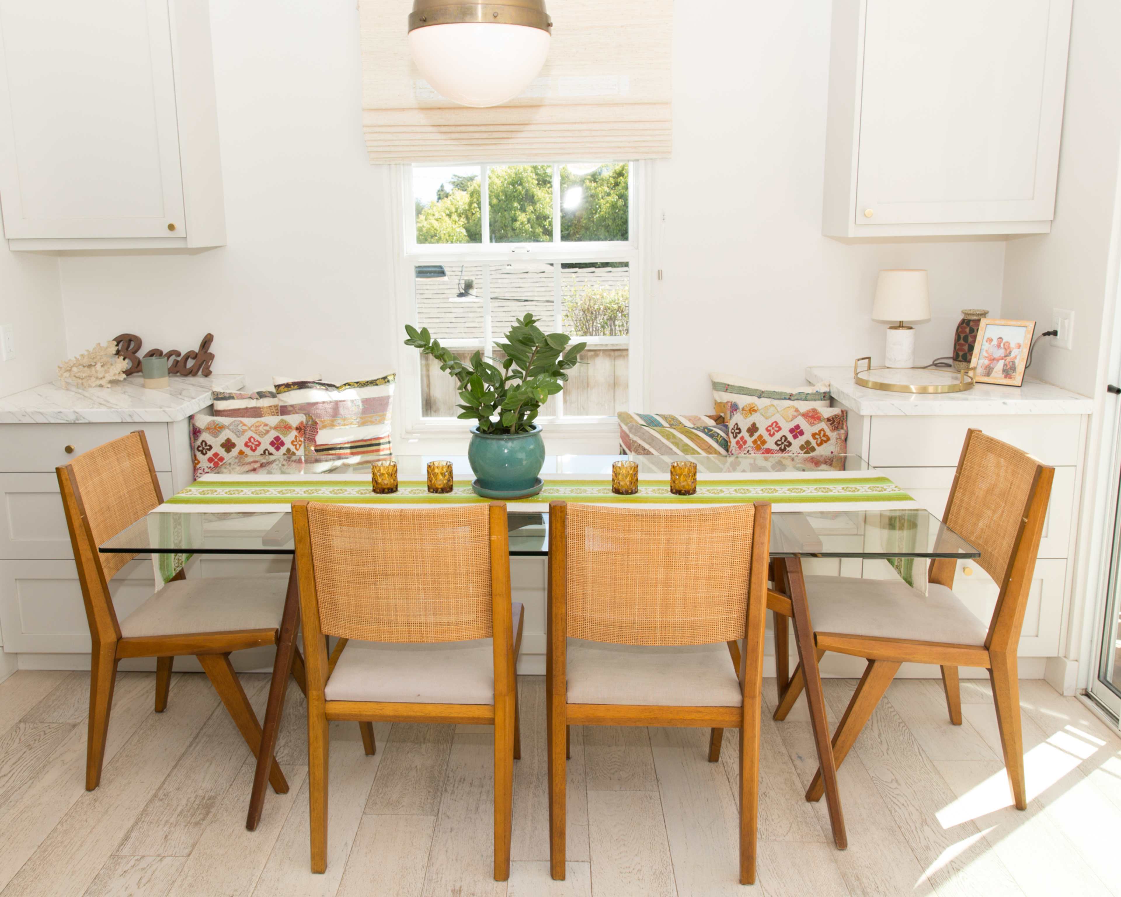 A glass dining table with four wooden chairs is set in a bright kitchen near a window, adorned with decorative items and a potted plant.
