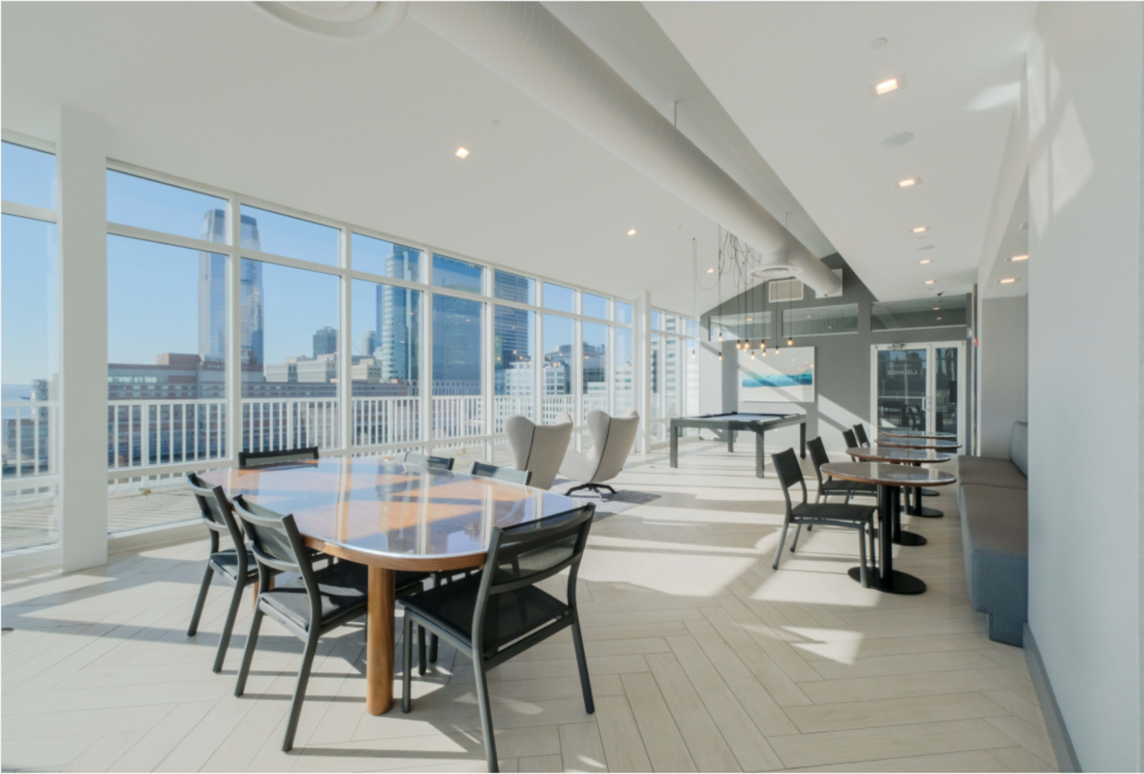 The image shows a well-lit modern lounge area with large windows, featuring a dining table, chairs, and a pool table, overlooking a city skyline.