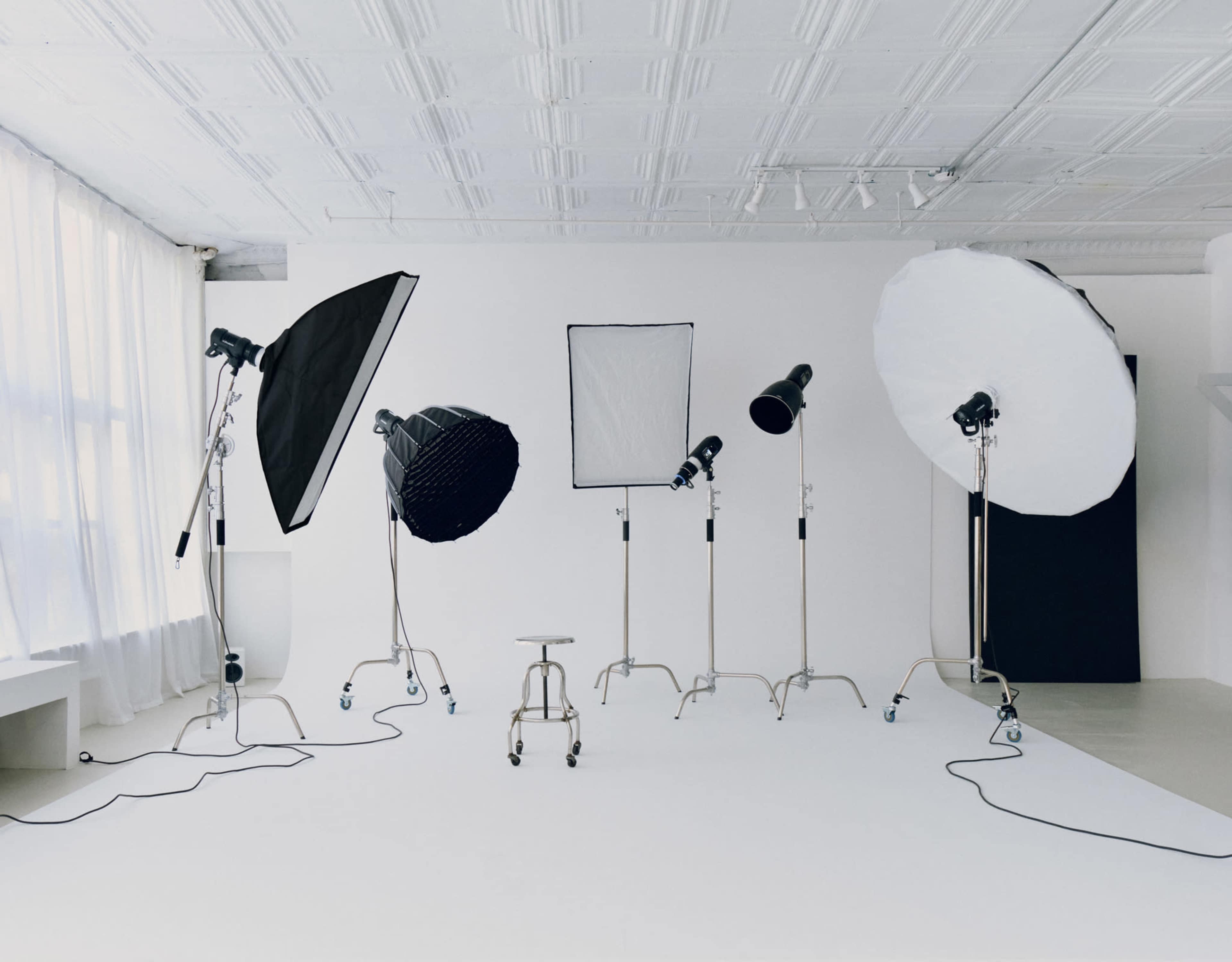 The image shows a photography studio equipped with various lighting setups, including softboxes and reflectors, arranged around a central empty stool.