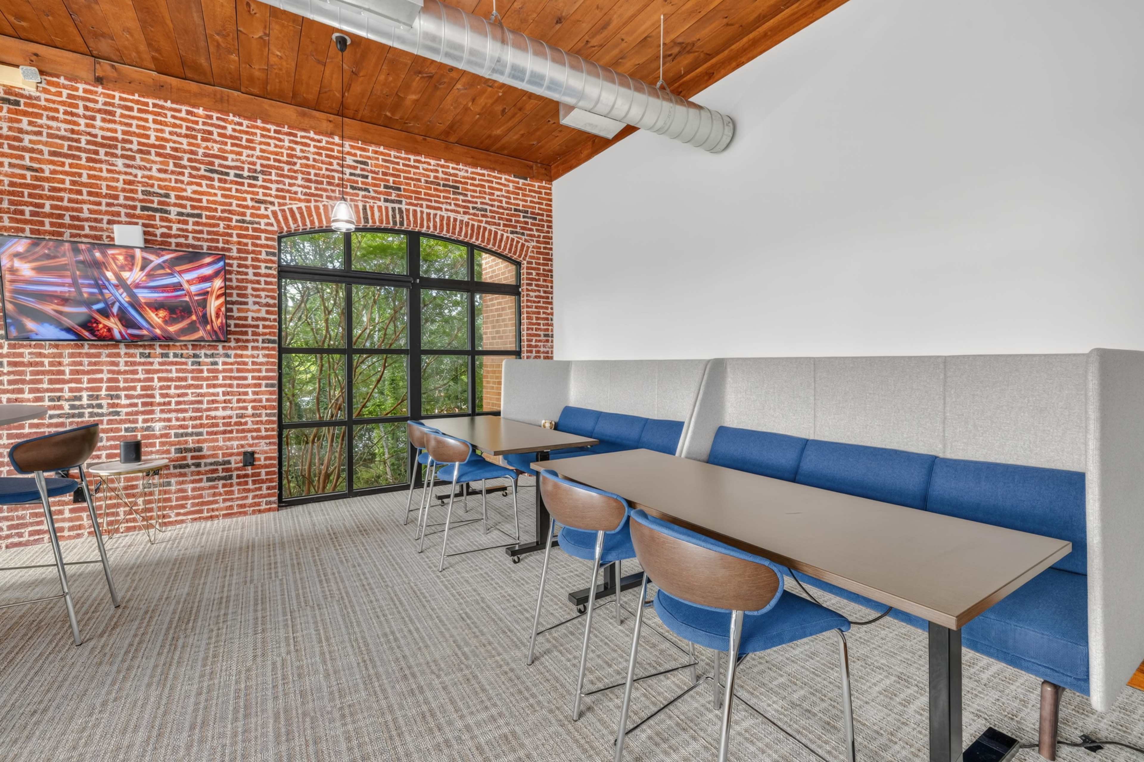 The image shows a modern meeting room with a brick wall, wooden ceiling, and several tables arranged with blue chairs, along with a wall-mounted television.