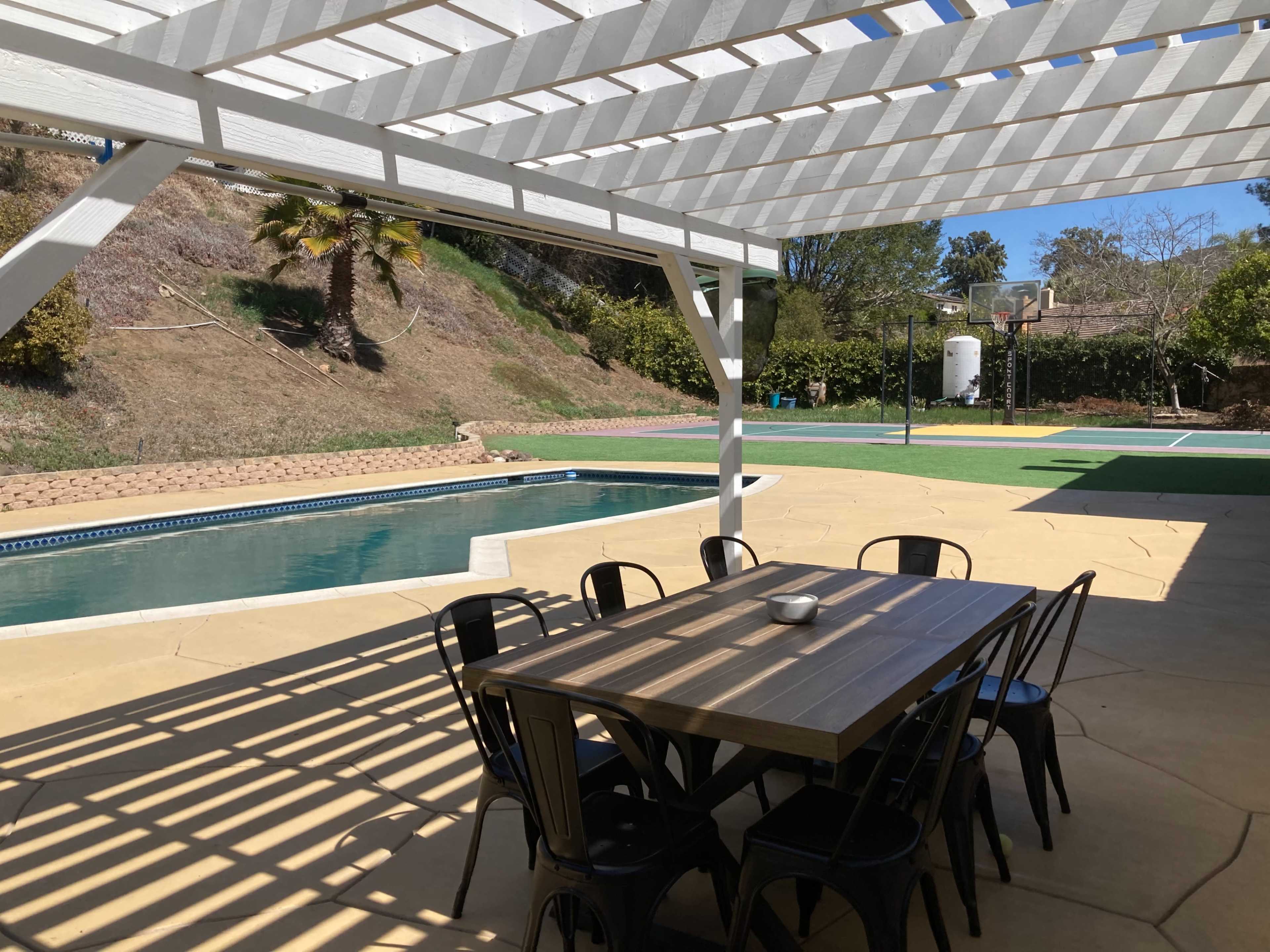 A backyard patio with a dining table and chairs beside a swimming pool and a basketball court in the background.