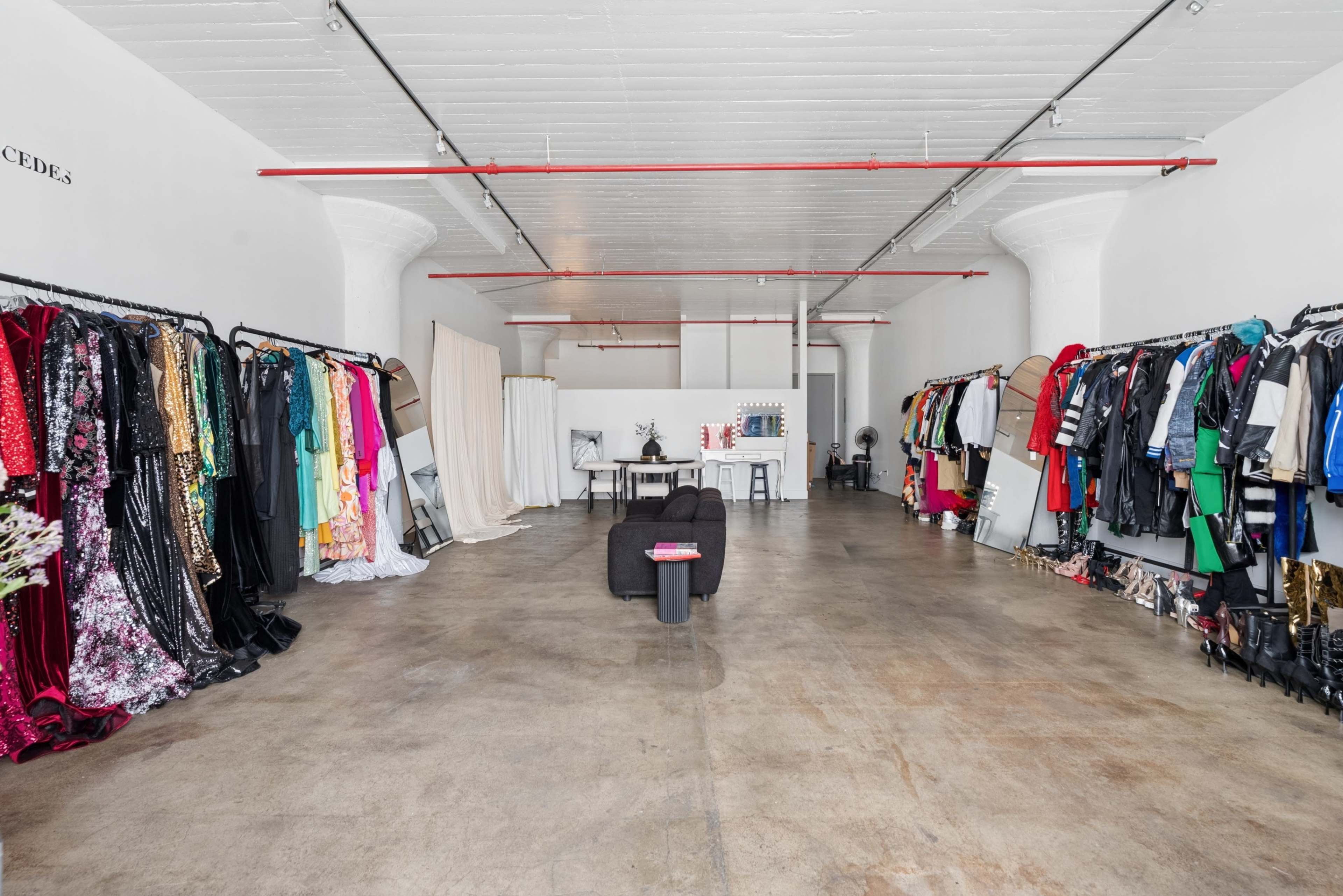 The image shows a spacious, well-lit clothing store or studio with racks of various garments on display along the walls and a table set in the center of the room.