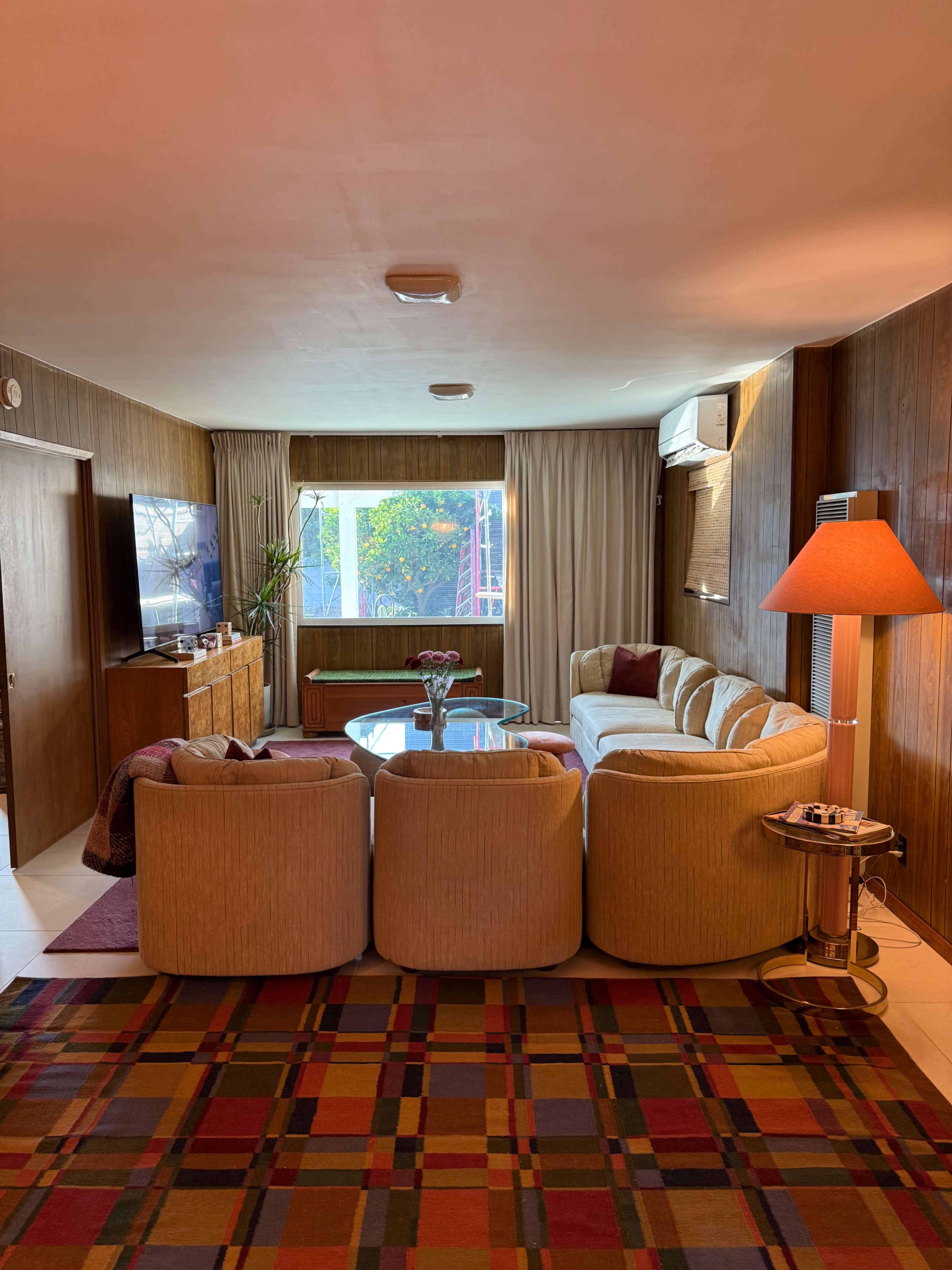 The image shows a cozy living room with a circular sofa, coffee table, and a lamp, featuring large windows that let in natural light.