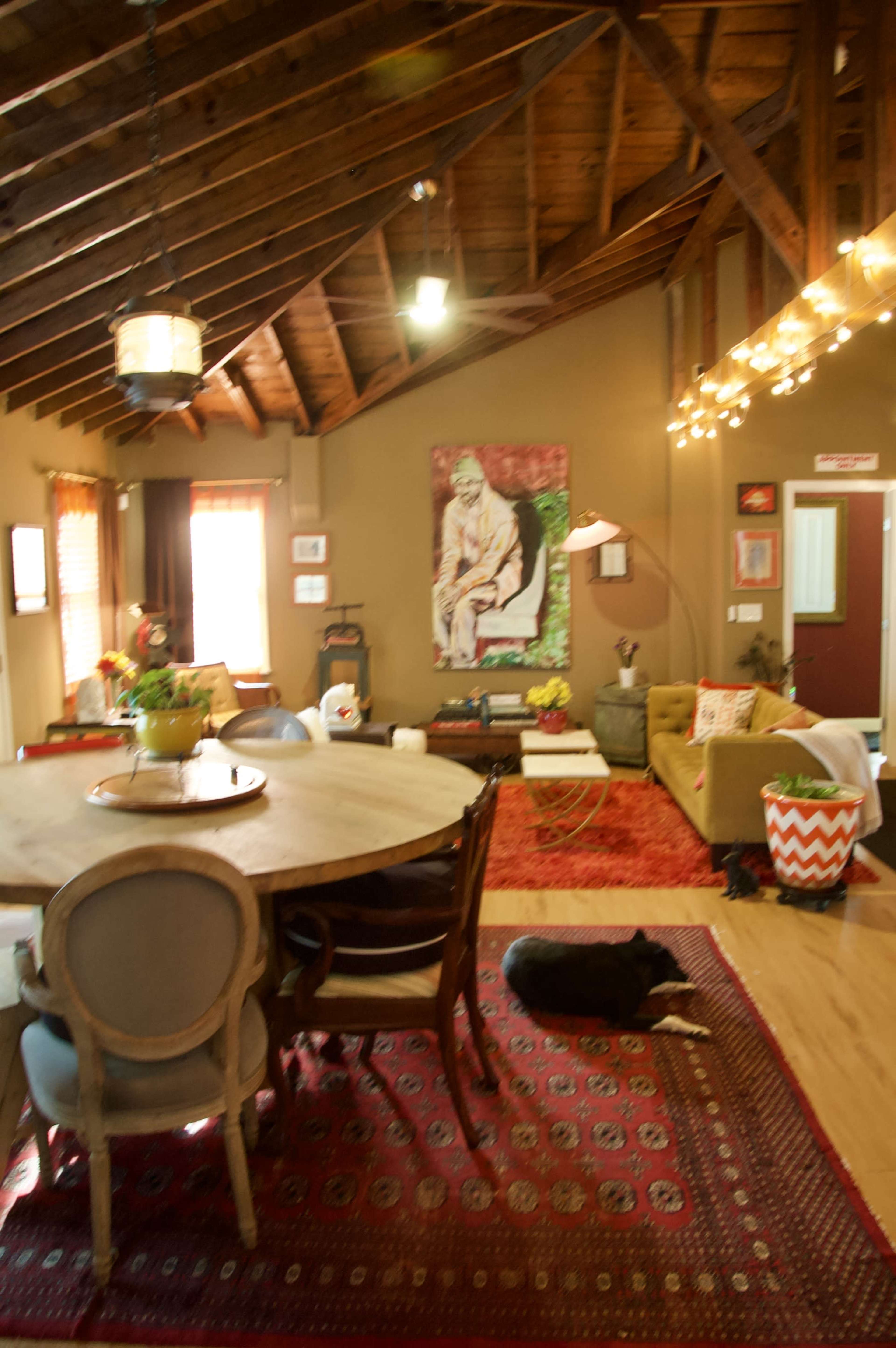 The image shows a cozy living room with a round wooden dining table, a variety of seating options, a colorful area rug, and a prominent wall art piece.